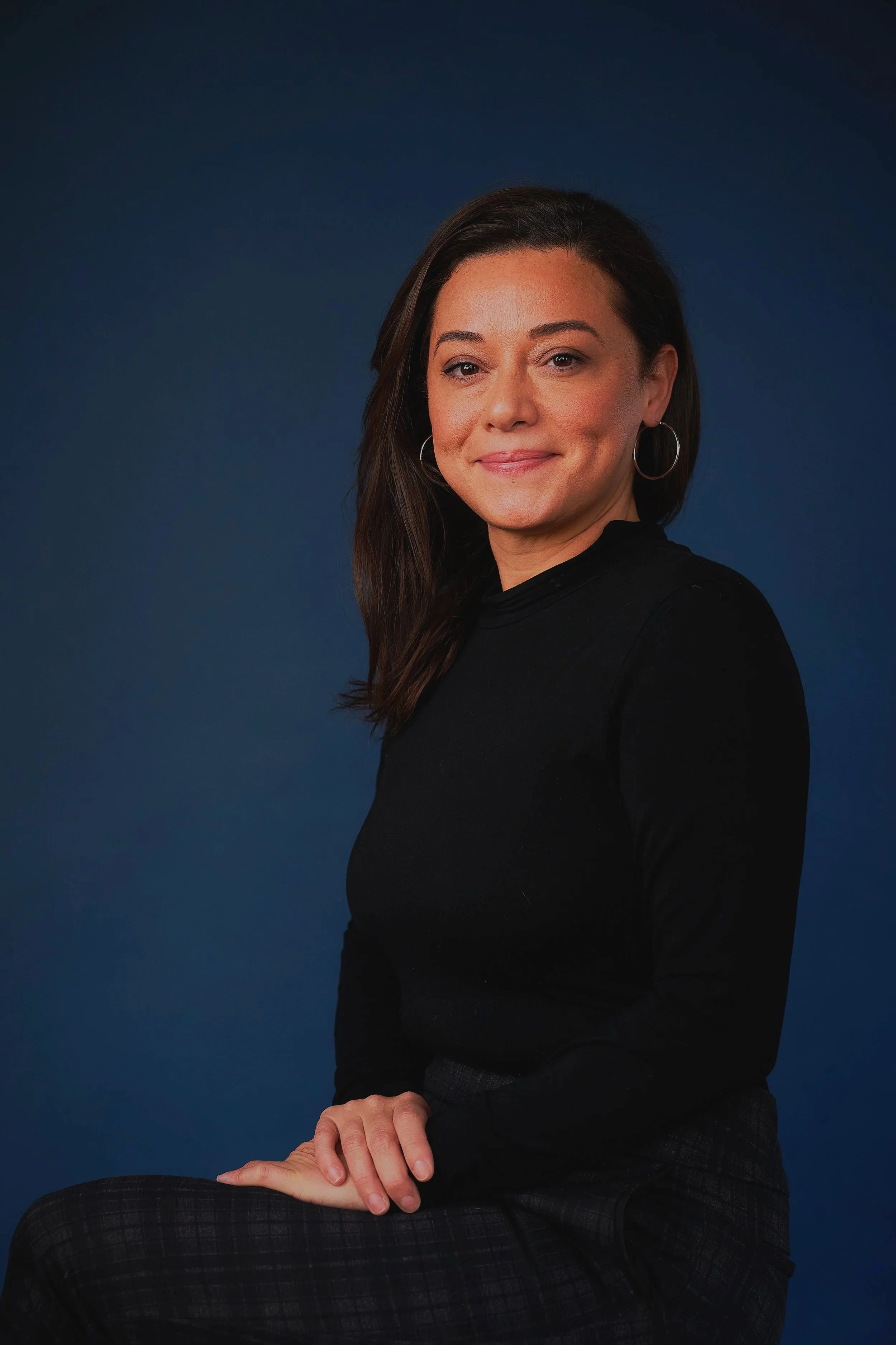 A woman with dark brown hair and hoop earrings, wearing a black top, sitting against a dark blue background.