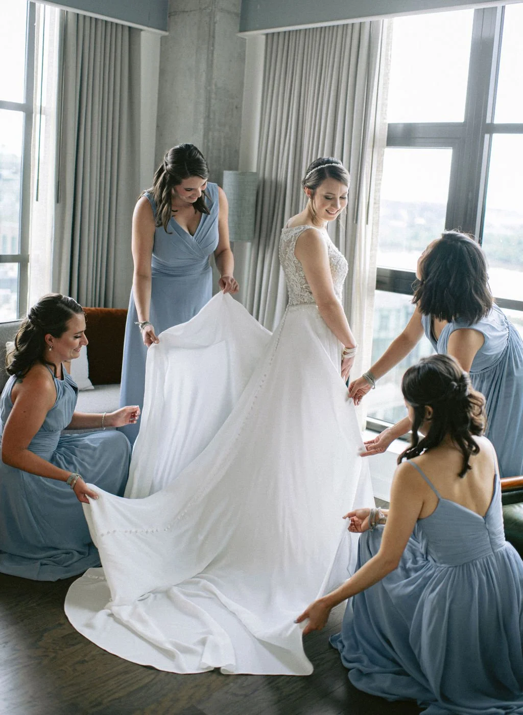 Bride in wedding dress surrounded by her bridesmaids adjusting her gown in a hotel room with large windows and curtains.