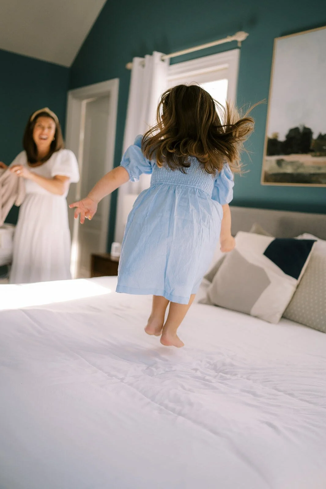 Young daughter jumping on a bed while her mother laughs during a playful, candid mother-daughter photo session
