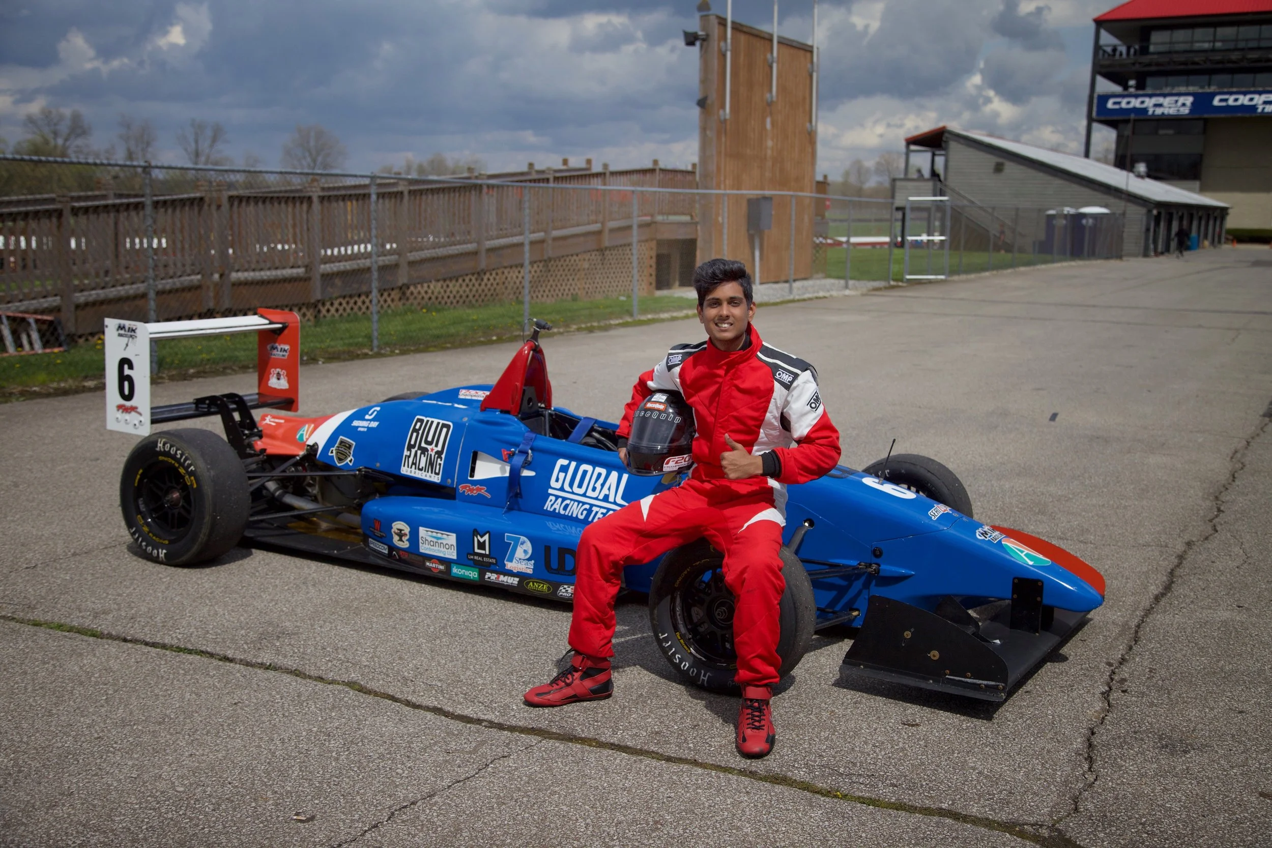 A young man in a red racing suit sitting on a blue open-wheel race car at a race track, giving a thumbs-up and smiling. The race car has various sponsor logos and the person is holding a black racing helmet.