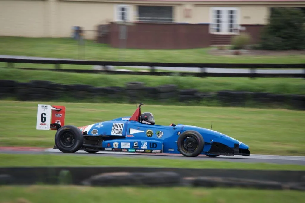 A blue race car with the number 6 is moving on a racetrack, with a driver wearing a black helmet, surrounded by green grass and a background of houses and a wooden fence.