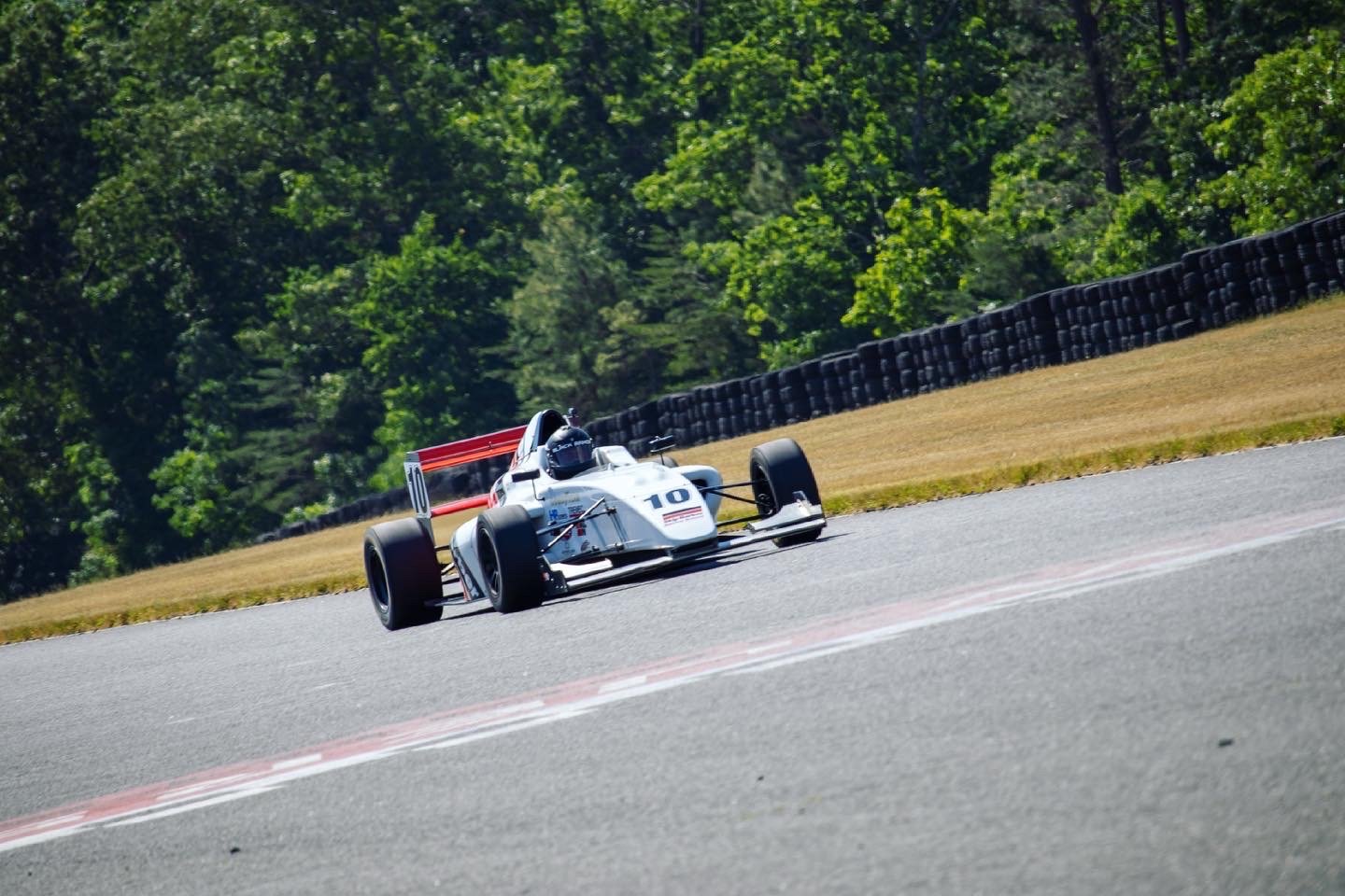 A vintage race car with the number 10 on it drives on a race track surrounded by greenery and tire barriers.