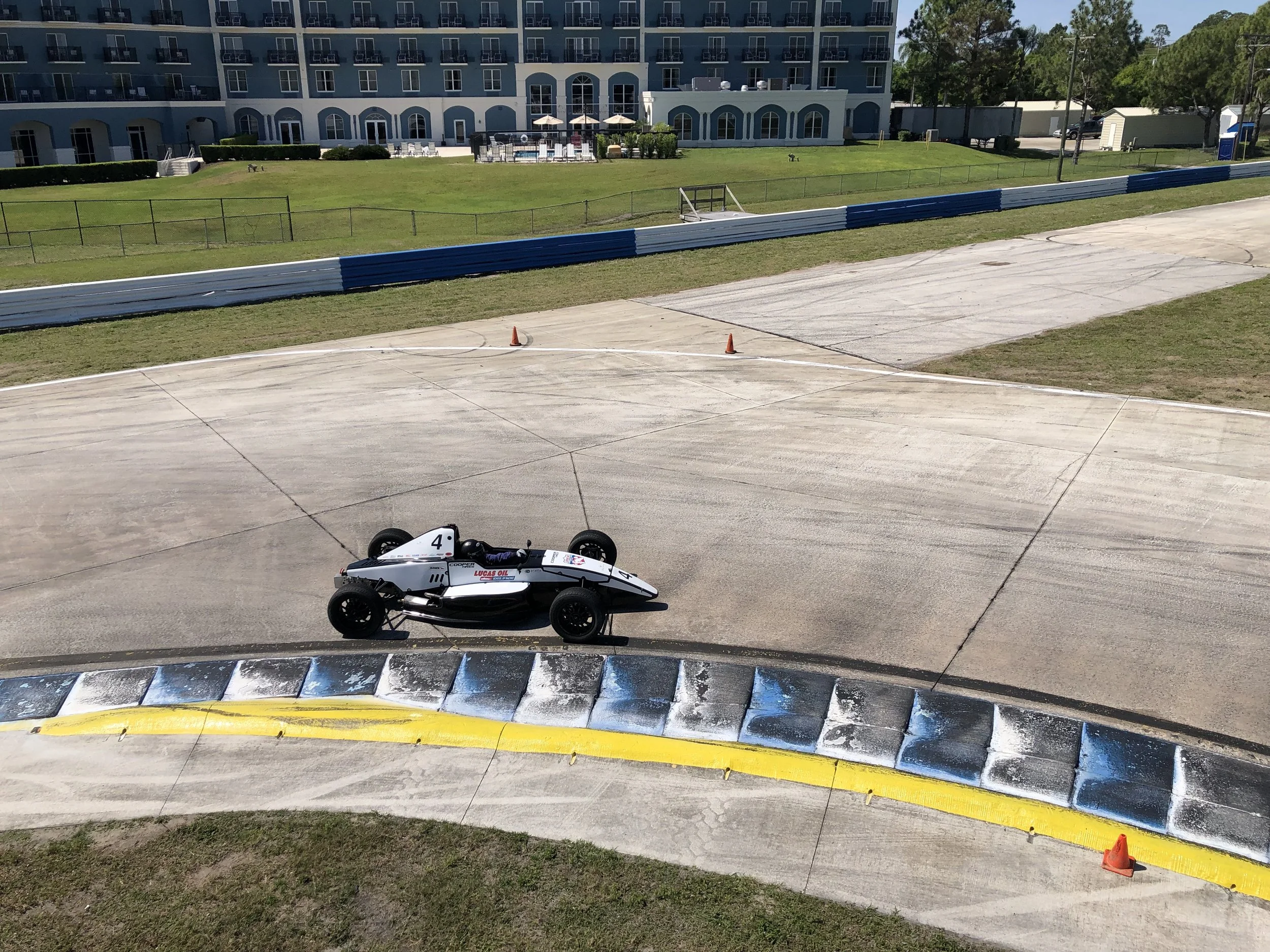 Image of a race car on a concrete track with colorful tire barriers in the foreground and a building with a pool and outdoor seating in the background.