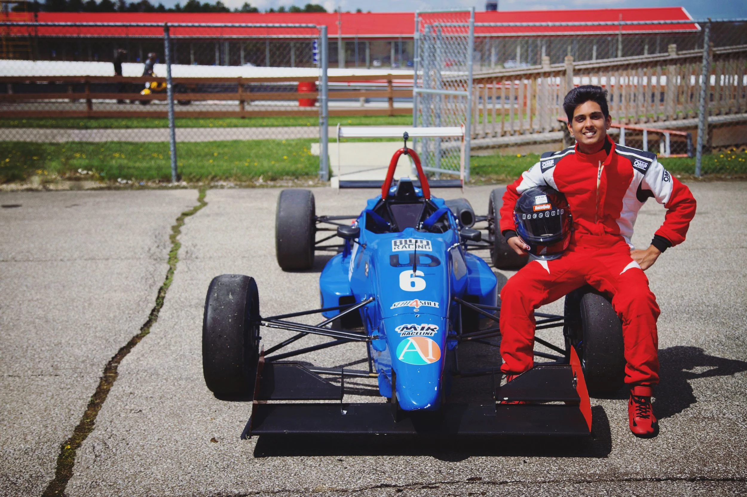 A smiling race car driver in a red racing suit and holding a helmet, standing next to a blue open-wheel race car with racing decals, at a race track.