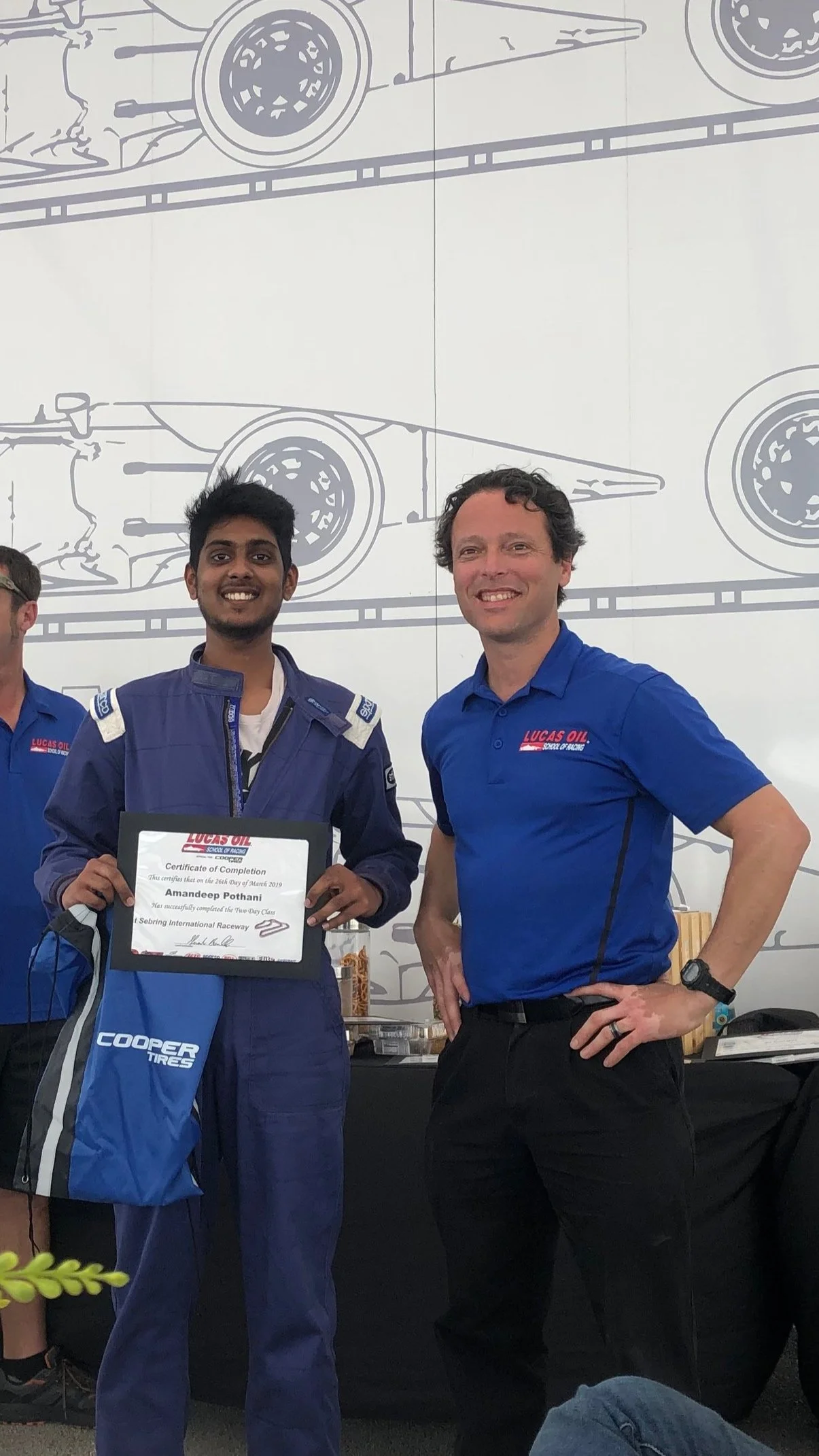 A young man in racing gear holding a certificate, standing next to a man in a blue polo shirt, both smiling, at an award ceremony with a backdrop of racing car illustrations.