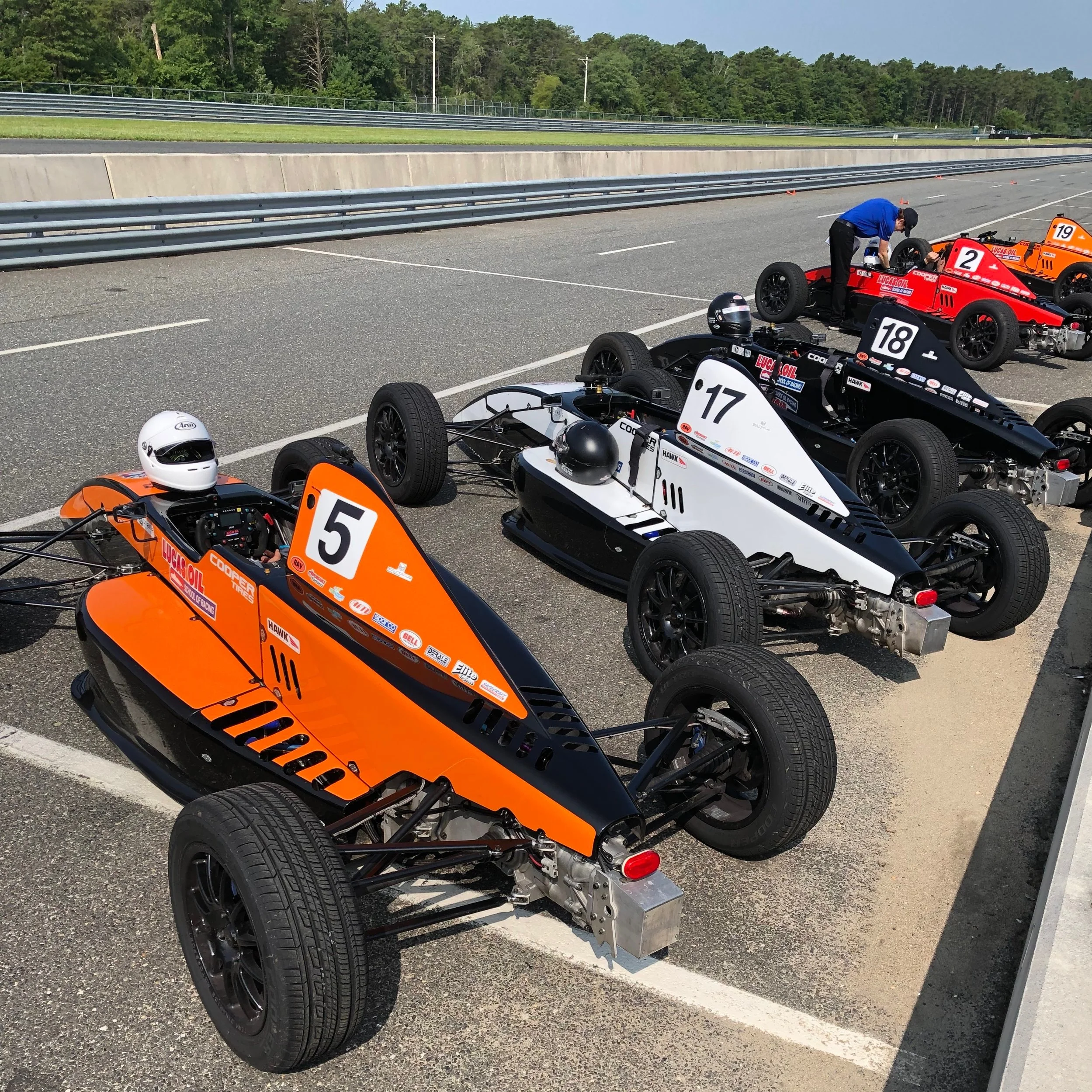 Race cars parked in a pit area with a person working on one of the cars, racing helmets on the ground, and a race track with trees in the background.