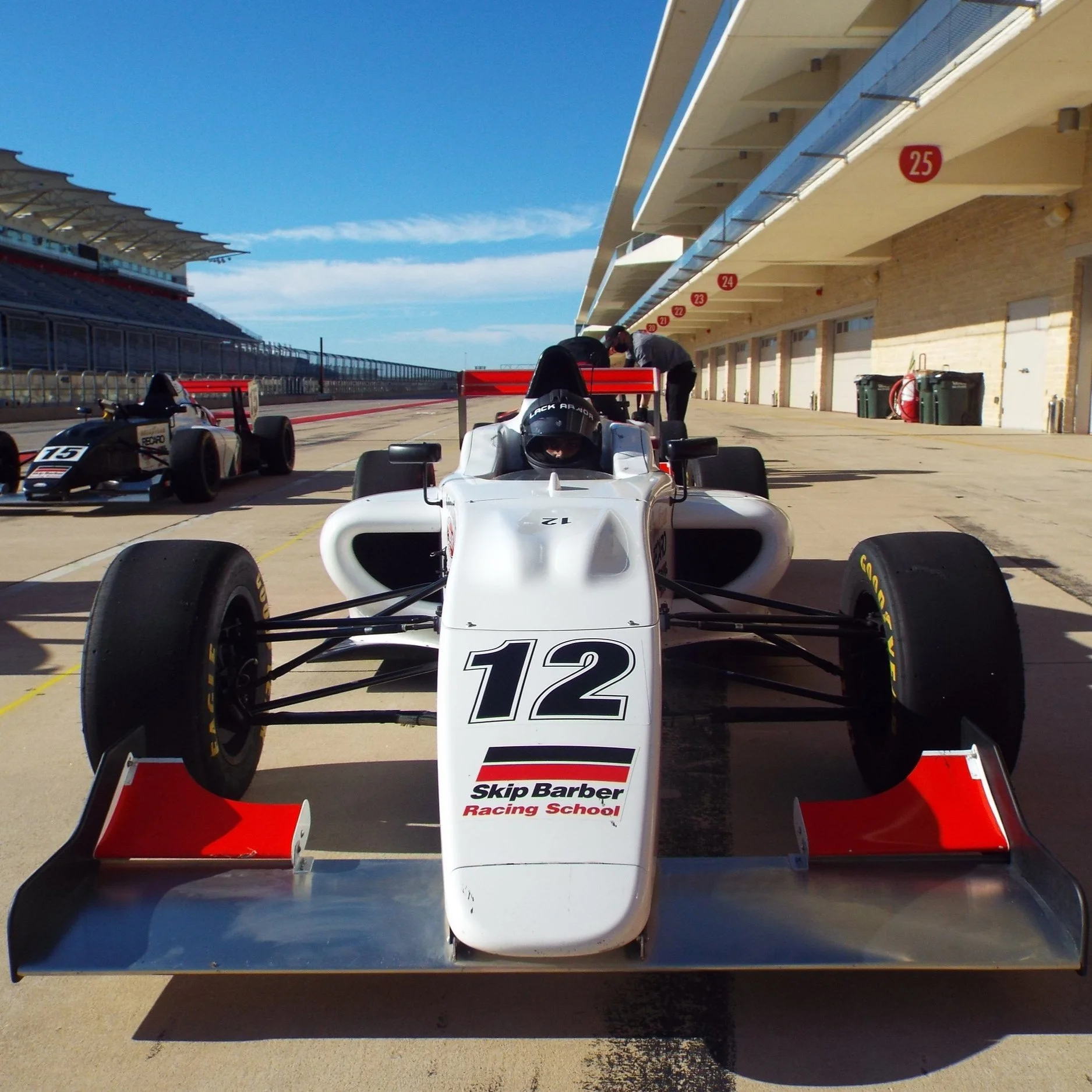 Open-wheel race car with the number 12 and Skip Barber Racing School logo parked in the pit lane at a racetrack. A technician is working on the car while another small open-wheel race car with the number 15 is nearby. The pit lane is under a building with numbered bays, and the sky is clear and blue.