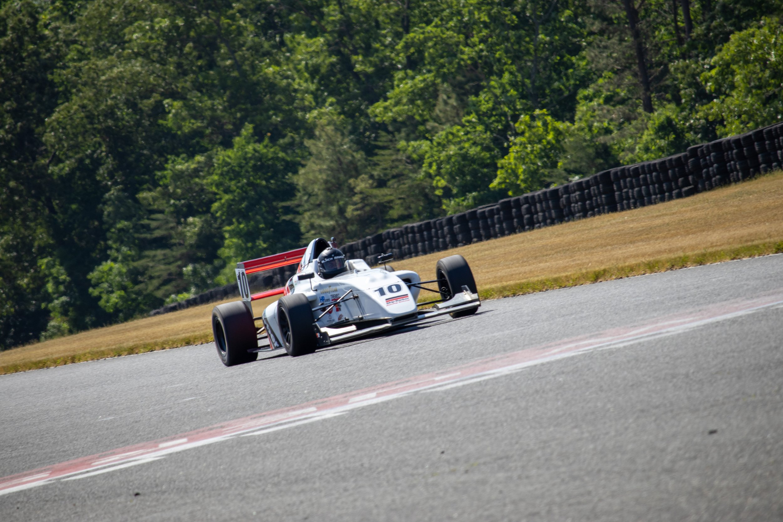 A white racing car with the number 10 on it, driving on a race track with green trees and tire barriers in the background.
