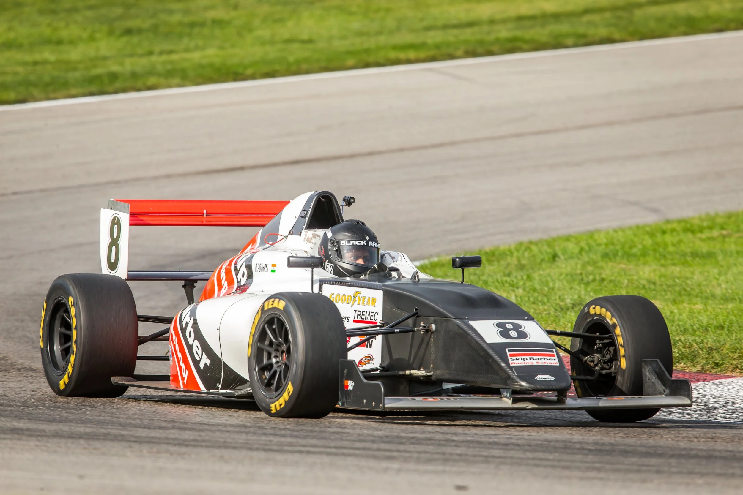 A race car on a track with a driver wearing a black helmet with 'Black Arm' written on it, and a red rear wing, competition decals, and Goodyear tires.