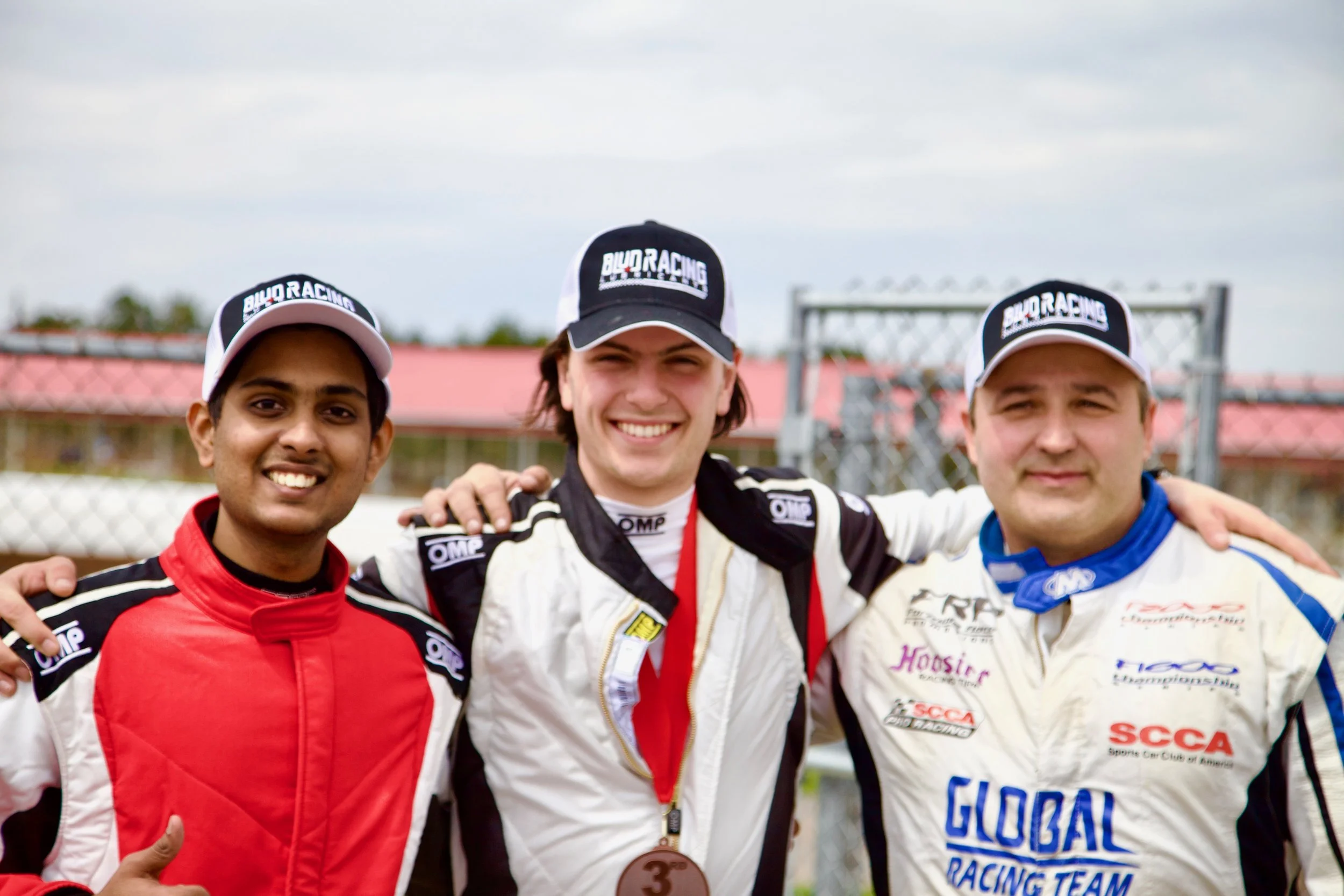 Three race car drivers standing together on a podium, smiling after a race, wearing racing suits and caps with 'BILD RACING' branding, with a chain-link fence and cloudy sky in the background.