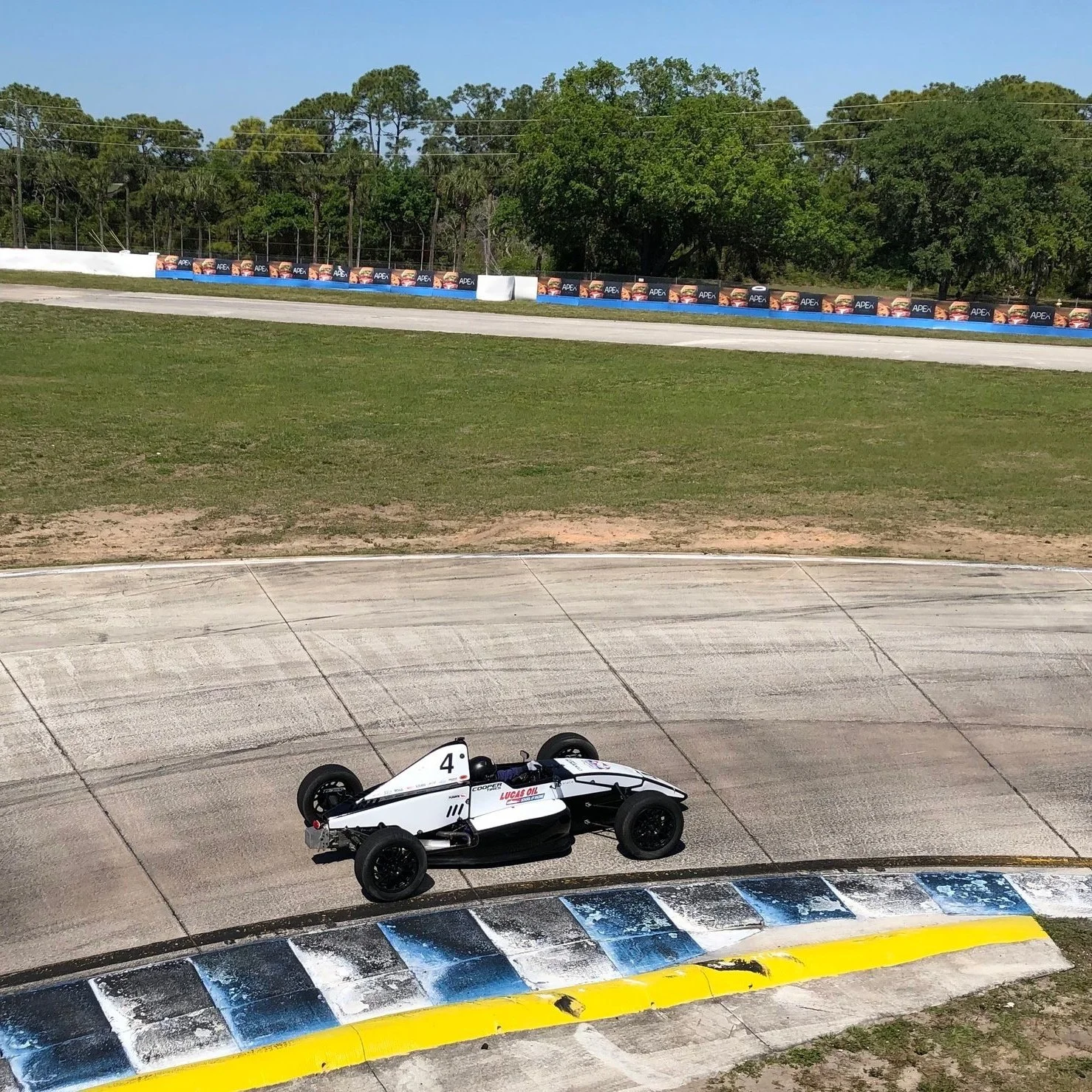 A white race car with the number 4 on its side on a race track, with a black and white checkered pattern on the curb in the foreground, and advertising banners with burgers and the word 'APEX' in the background.