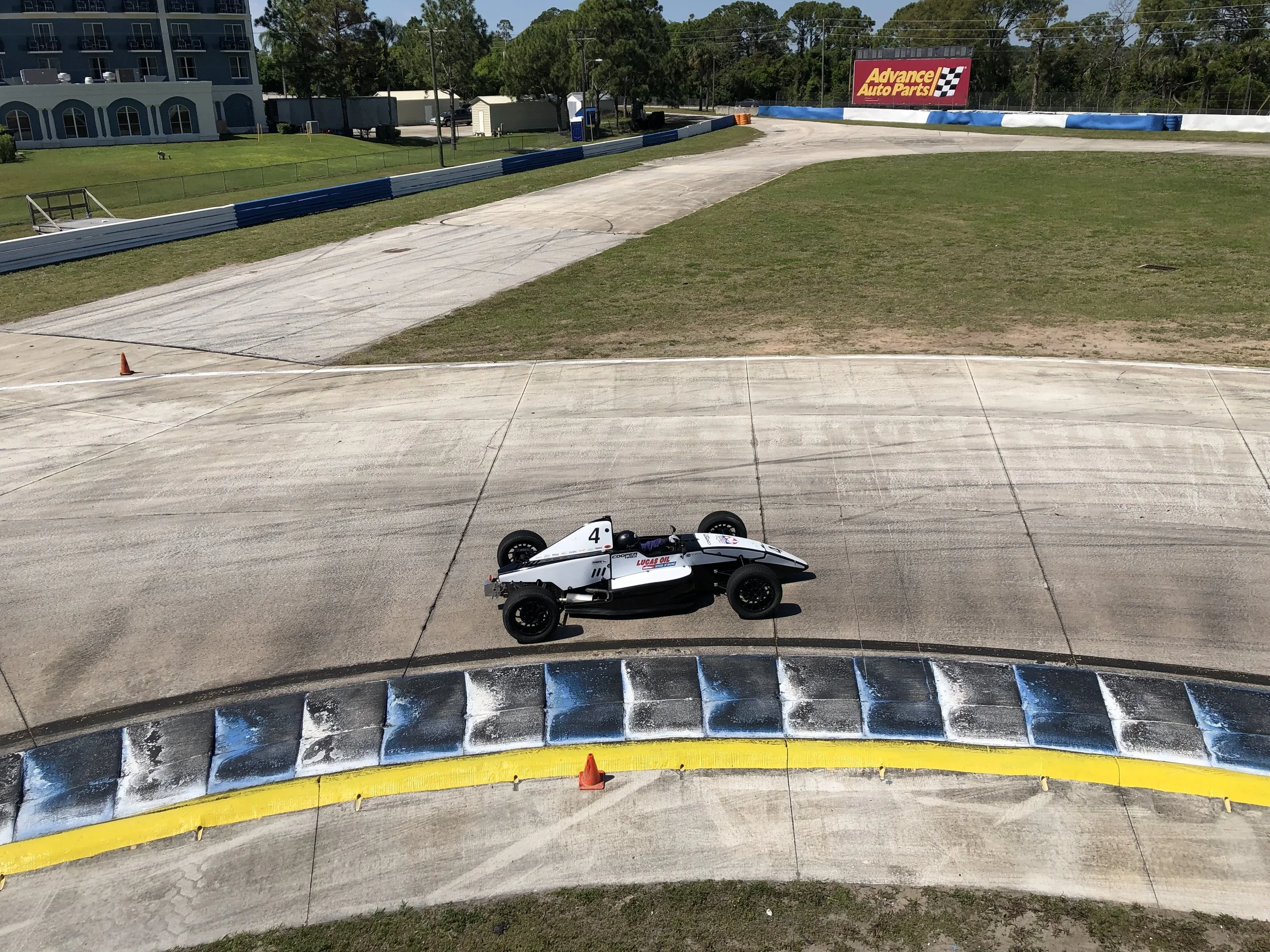 A go-kart on a race track with a curving barrier wall, a construction cone, and a sign for Advance Auto Parts at the finish line.