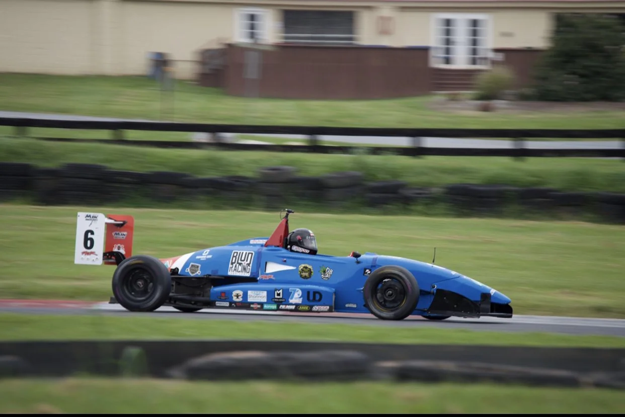 A blue racing car with the number 6 on a white sign, speeding on a race track with black tire barriers and a fencing in the background.