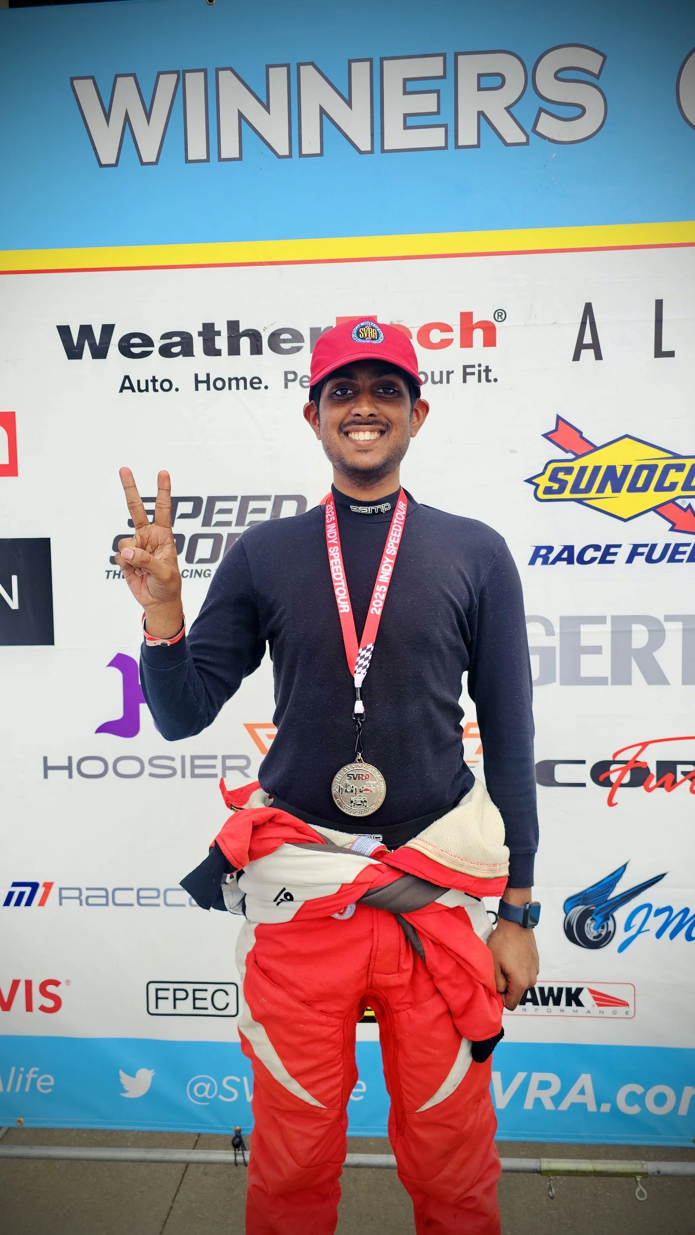 A man in racing attire standing in front of a backdrop with logos, smiling, holding up a peace sign, wearing a red cap, a medal around his neck, and red racing pants.