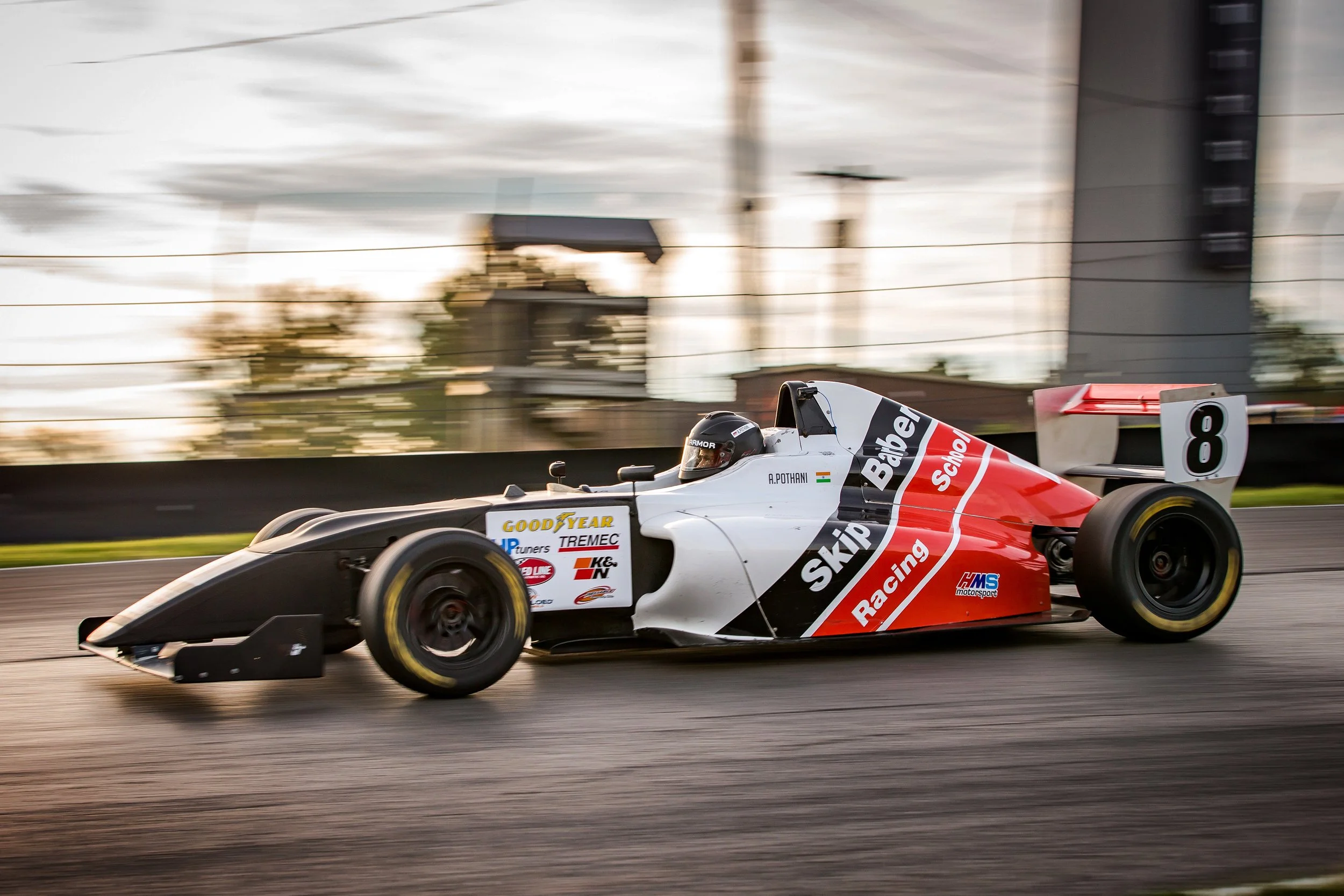 A white Formula race car with red and black accents speeds along a track, with motion blur in the background, and displays sponsorship logos including Skip Raceing, Goodyear, and others.