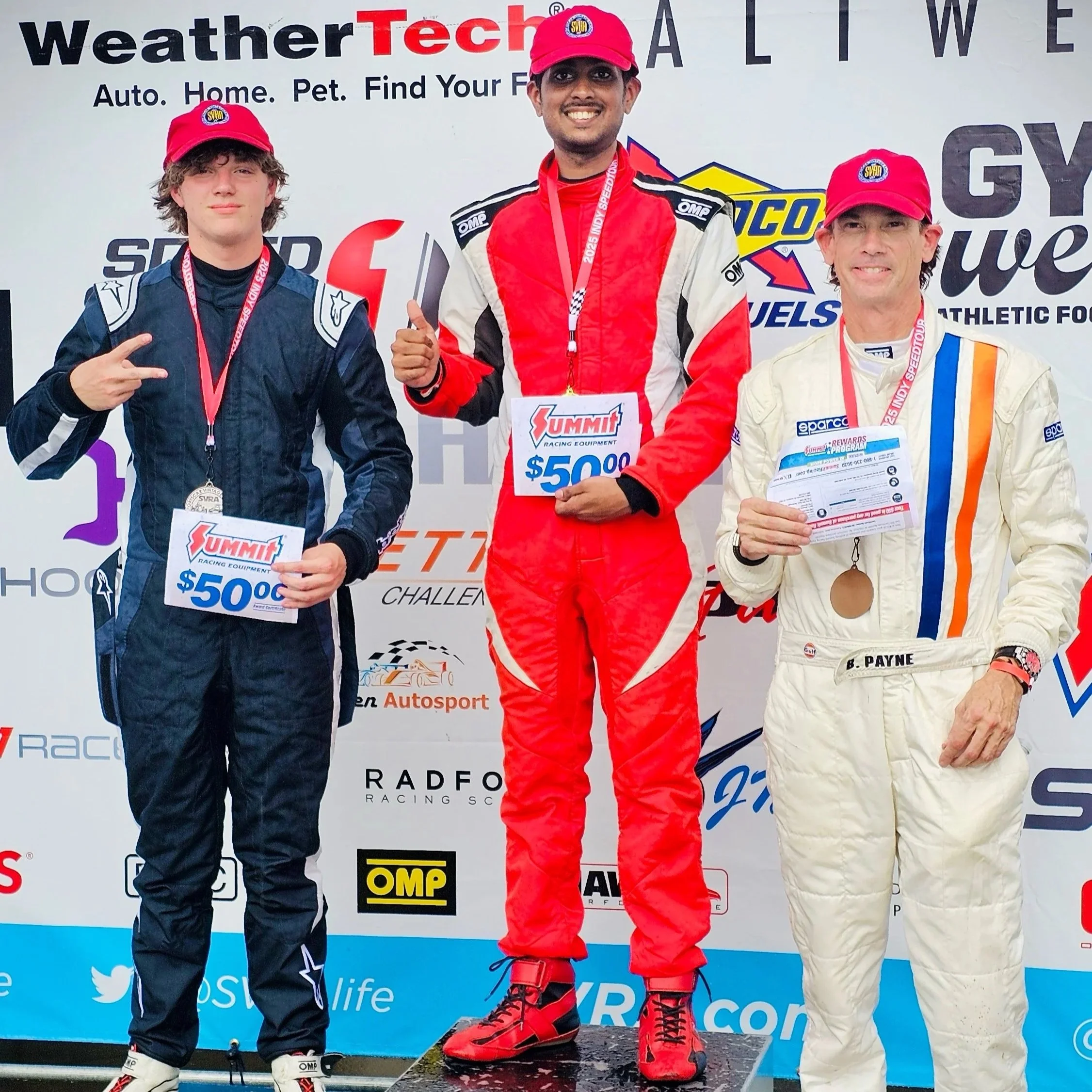 Three race car drivers standing on a winners' podium, wearing racing suits and red caps, holding certificates and medals, smiling. Behind them is a backdrop with various sponsor logos and text.