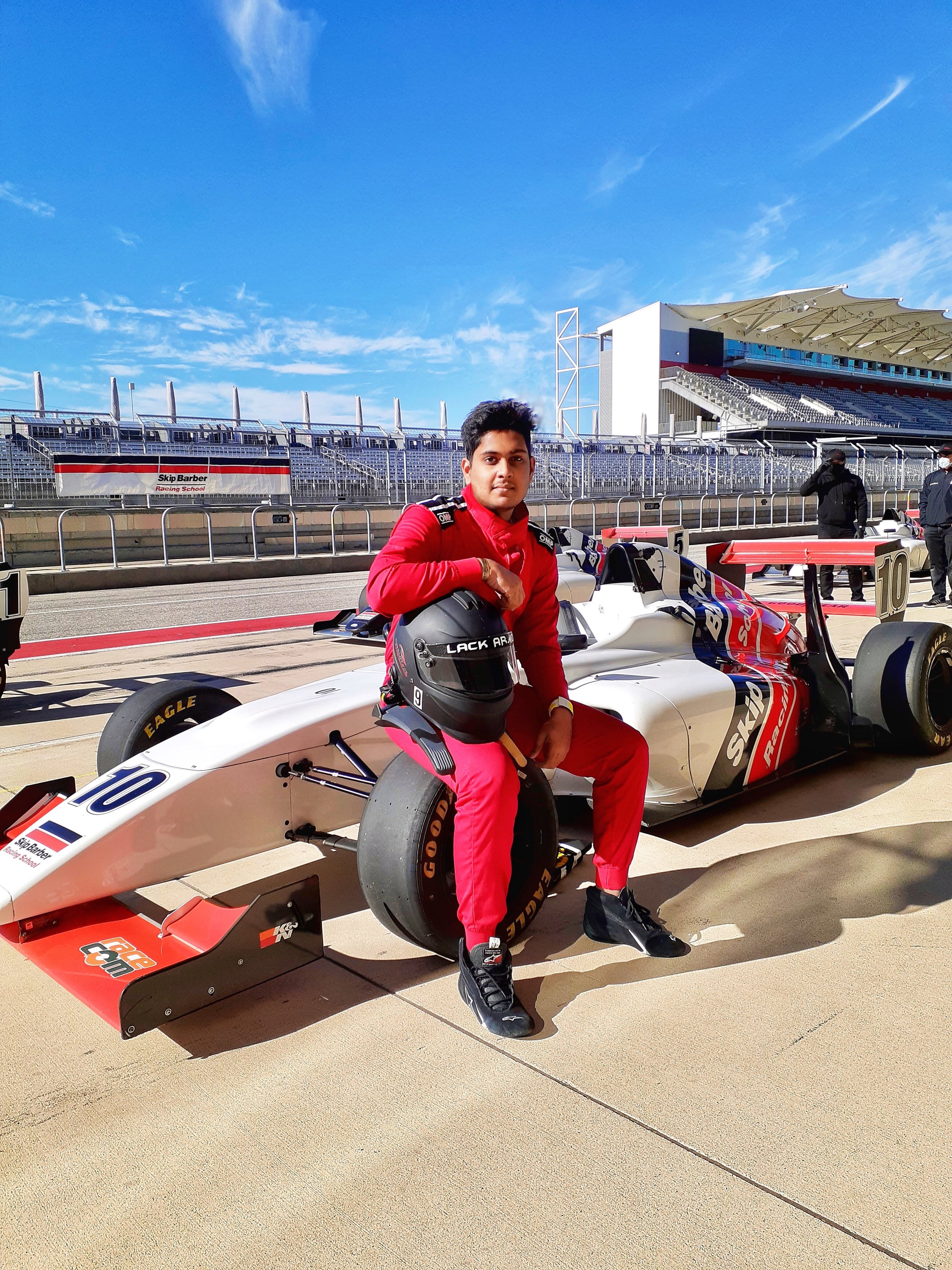 A young man in racing gear sitting on a race car at a racing track with empty grandstands and a clear blue sky in the background.