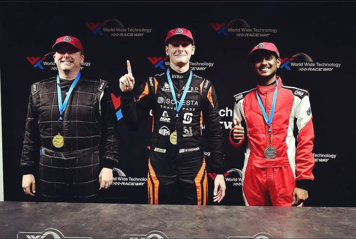 Three race car drivers standing on a podium at the World Wide Technology Raceway, wearing racing suits and medals, with a black backdrop displaying the raceway logo.