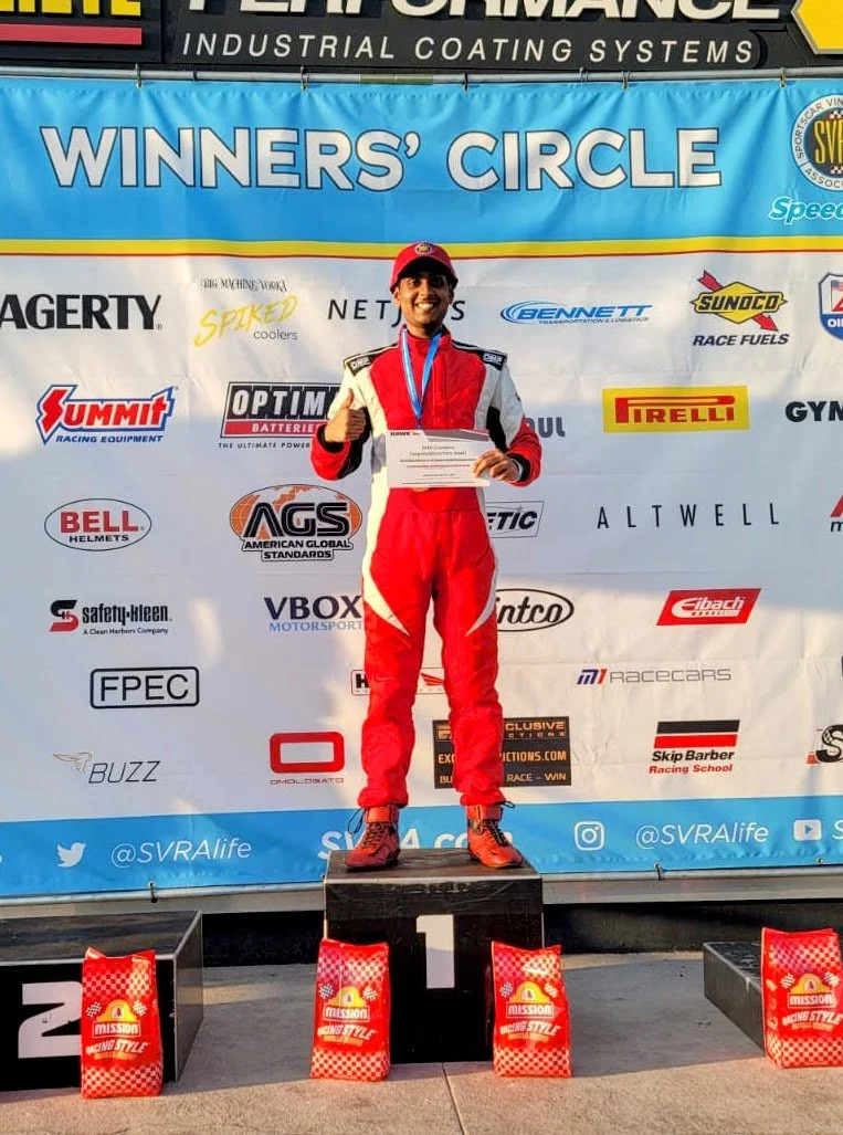 A race car driver in a red racing suit and cap standing on a winners' podium, holding a certificate, with a medal around his neck, at an awards ceremony. The backdrop features the words 'WINNERS' CIRCLE' and various sponsor logos.