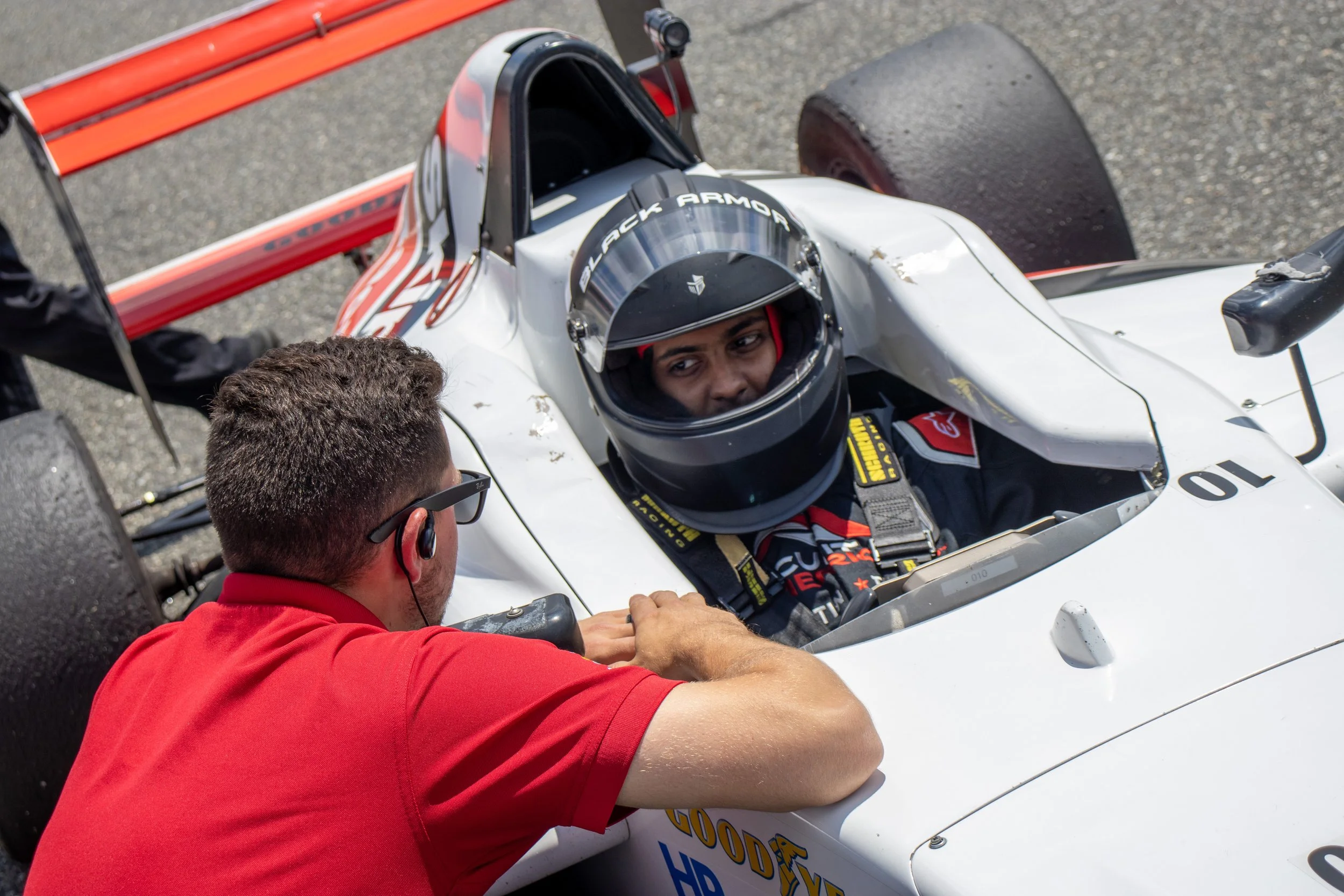 Race car driver in a white race car, wearing a black helmet with a visor, seated in the car, and a crew member in a red shirt working on the car's front.