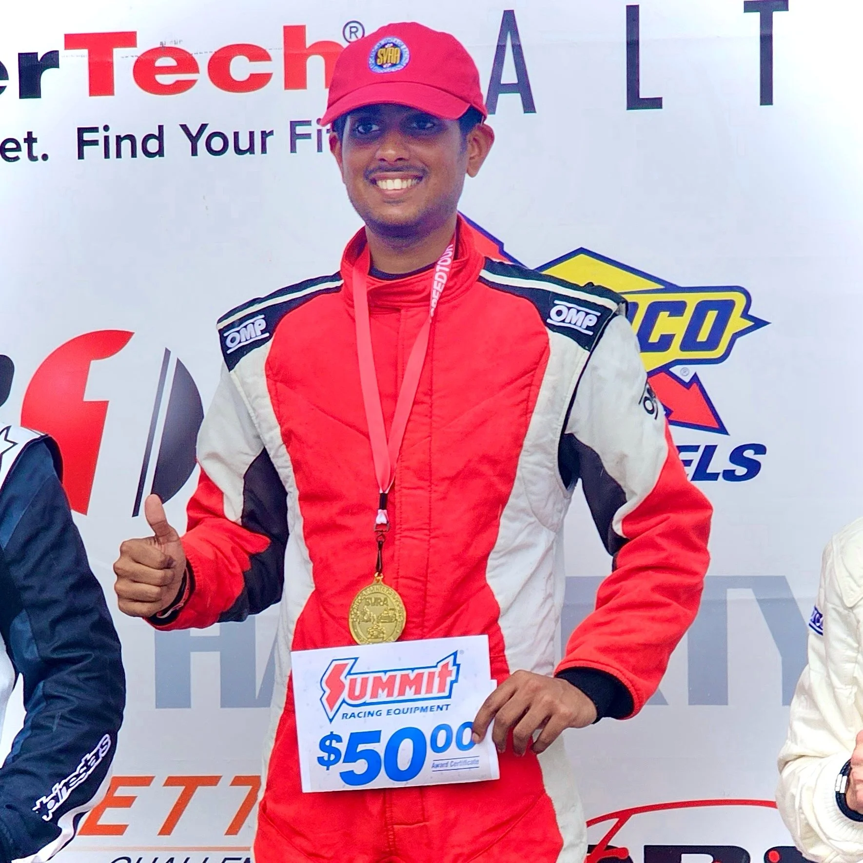 A young man in a red racing suit and red cap smiling and giving a thumbs-up, wearing a gold medal around his neck, at a racing event. He is holding a sign that reads there is a $50,000 prize. The background features a banner with logos and the words 