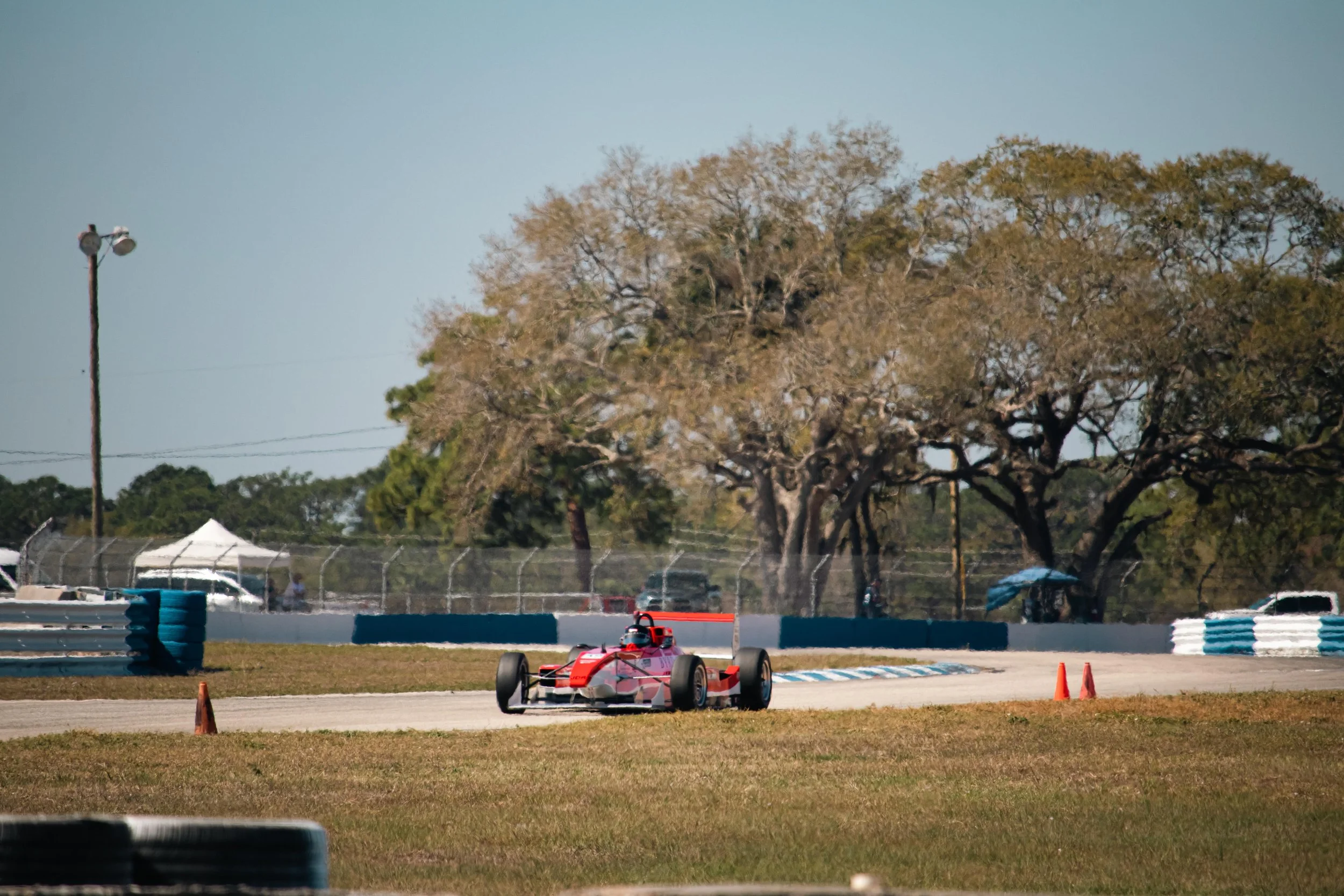 A red race car on a race track with orange cones, barrier walls, and large trees in the background.