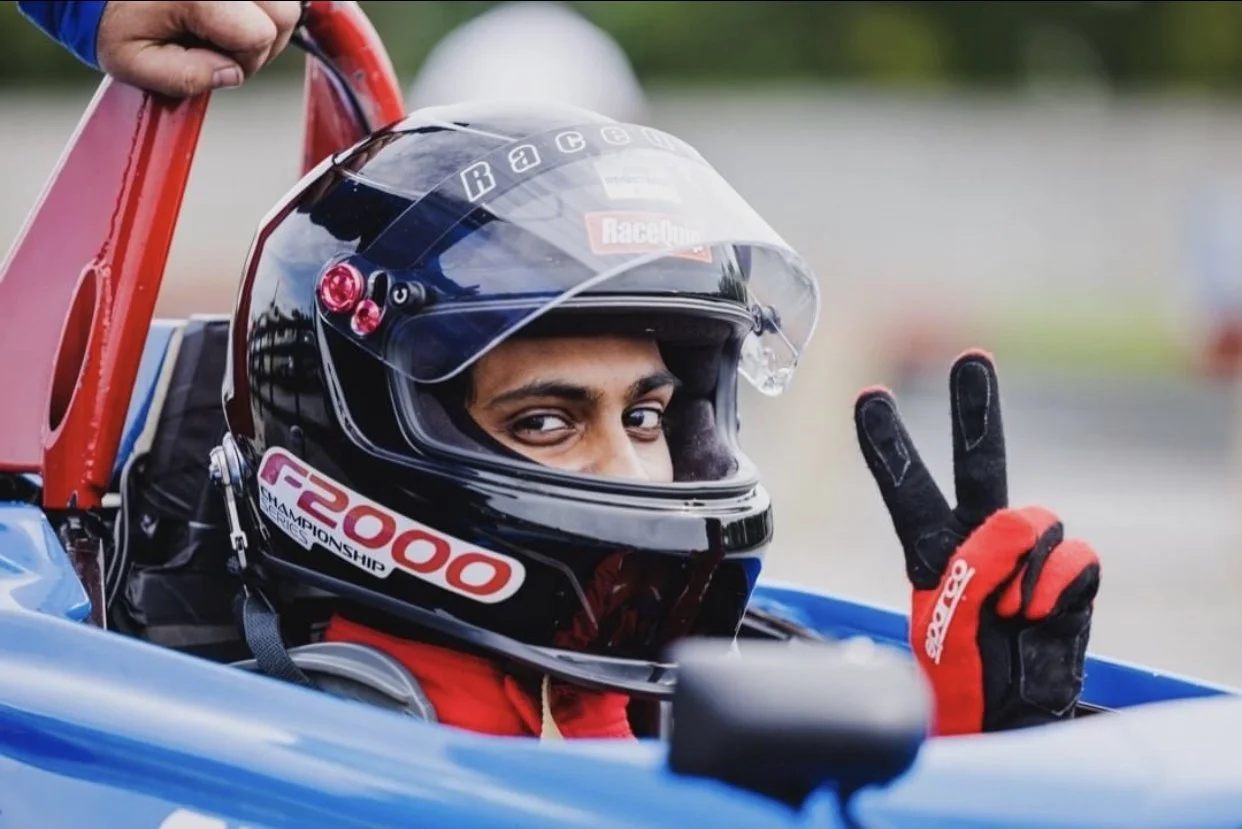 A race car driver wearing a helmet and red gloves, sitting in a blue race car, making a peace sign with his hand.