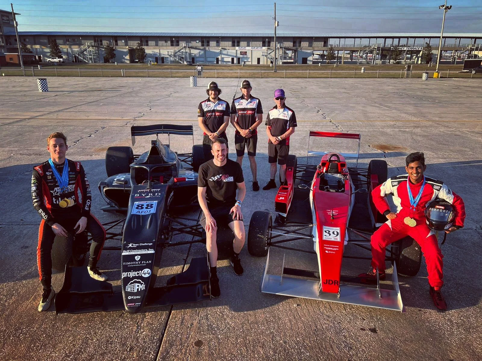 Six people, three in racing suits and three in casual clothes, pose with two open-wheel race cars on an outdoor racetrack. The person on the left in a racing suit holds a helmet with medals around his neck. The person in the center in casual clothes sits on the front of a black race car. The person on the right in a racing suit holds a helmet with medals hanging around his neck. The two race cars are black and red, with racing numbers 88 and 39 respectively.