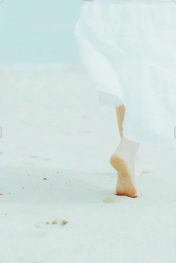 Close-up of a person walking barefoot on white sand at the beach, with part of their white dress visible.
