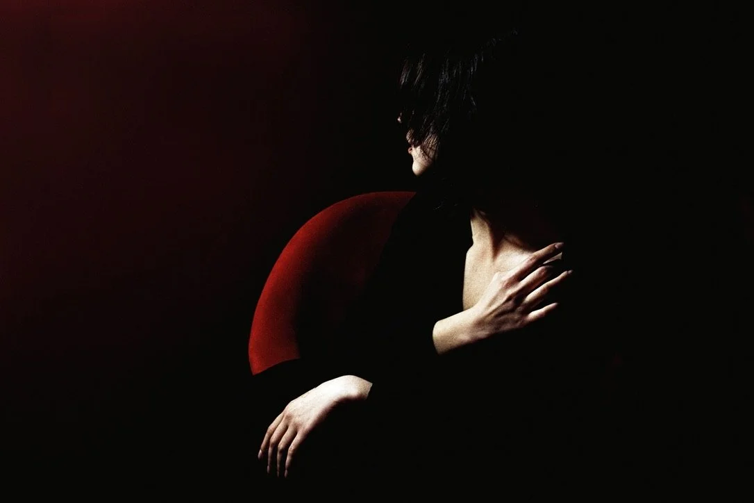 A person sitting on a dark red chair, hugging their knees and covering their face, with hair partially covering their face, set against a black background.