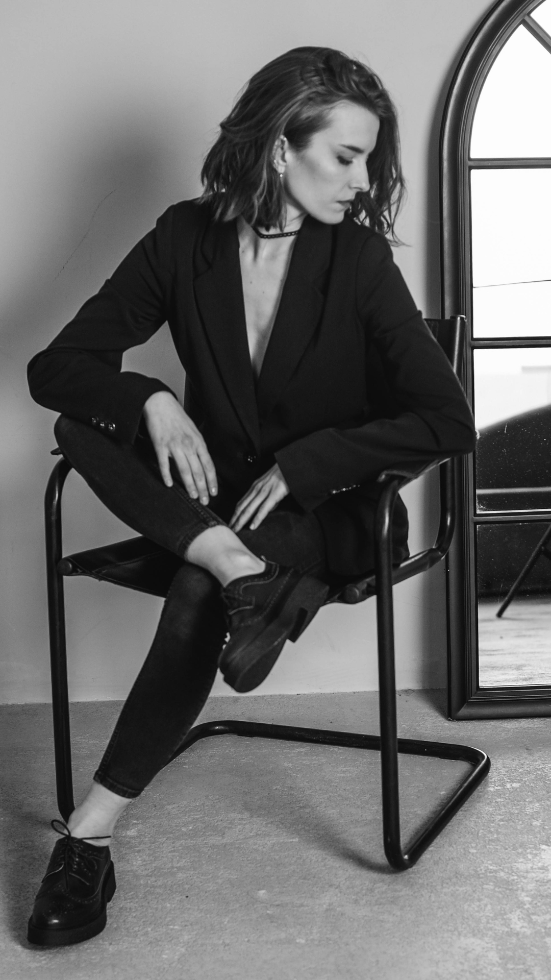 A woman with shoulder-length hair, dressed in a black blazer and dark jeans, seated on a black chair with one leg crossed over the other, near a mirror and a window, in a black and white photo.
