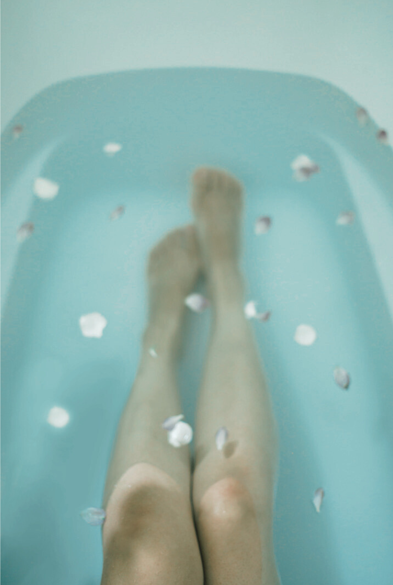Lying legs partially submerged in a blue bathtub with floating flower petals.