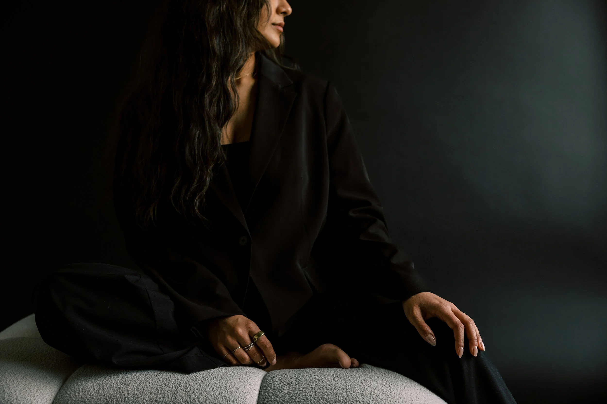 Woman sitting on a light gray textured bench against a dark background, dressed in black with long curly hair, looking to the side.