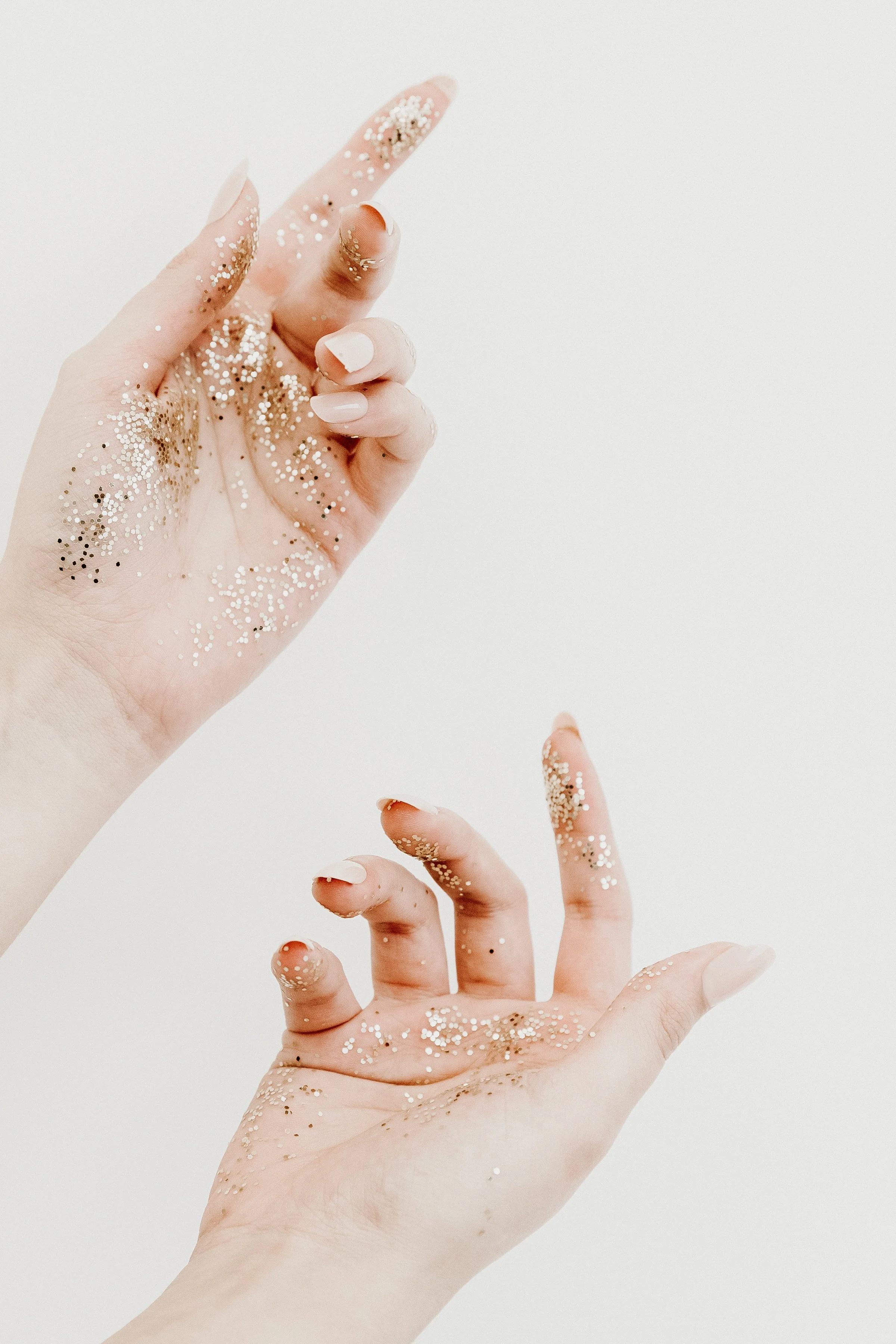 Two hands, one with light skin and painted nails, the other with darker skin, are decorated with white and gold glitter. The hands are positioned against a plain white background.