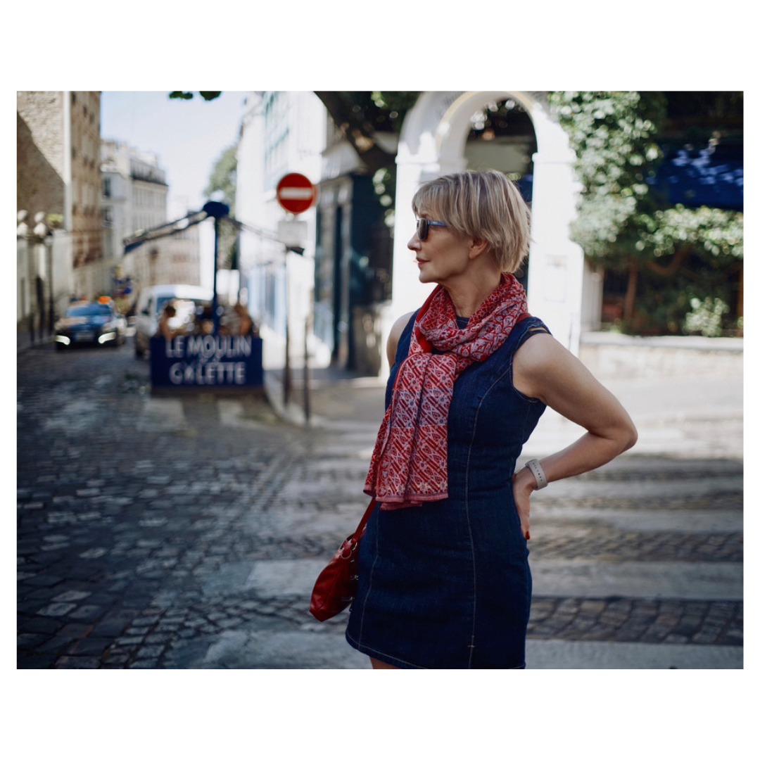 A woman with blonde hair wearing sunglasses, a denim dress, and a red patterned scarf stands on a cobblestone street in Paris, France, with her hand on her hip, looking to the side. In the background, there is a sign for a restaurant or cafe named Le Moulin Cleette, and some cars and buildings are visible.