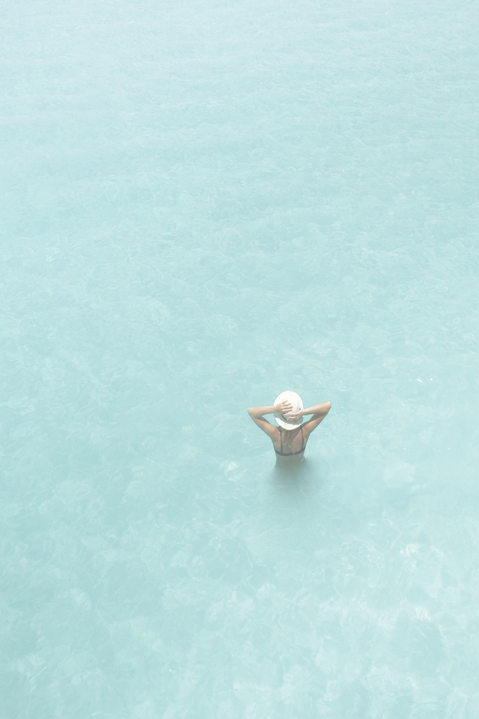 Top-down view of a woman standing in a pool, wearing a white wide-brimmed hat and black swimwear.