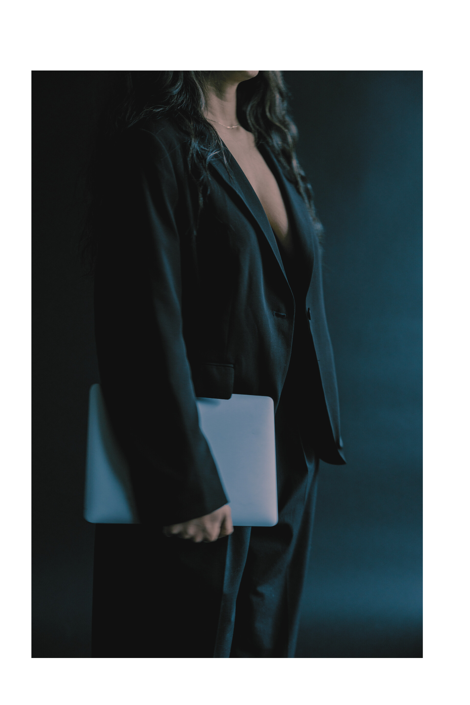 A woman in a black suit holding a laptop, standing in front of a dark background.