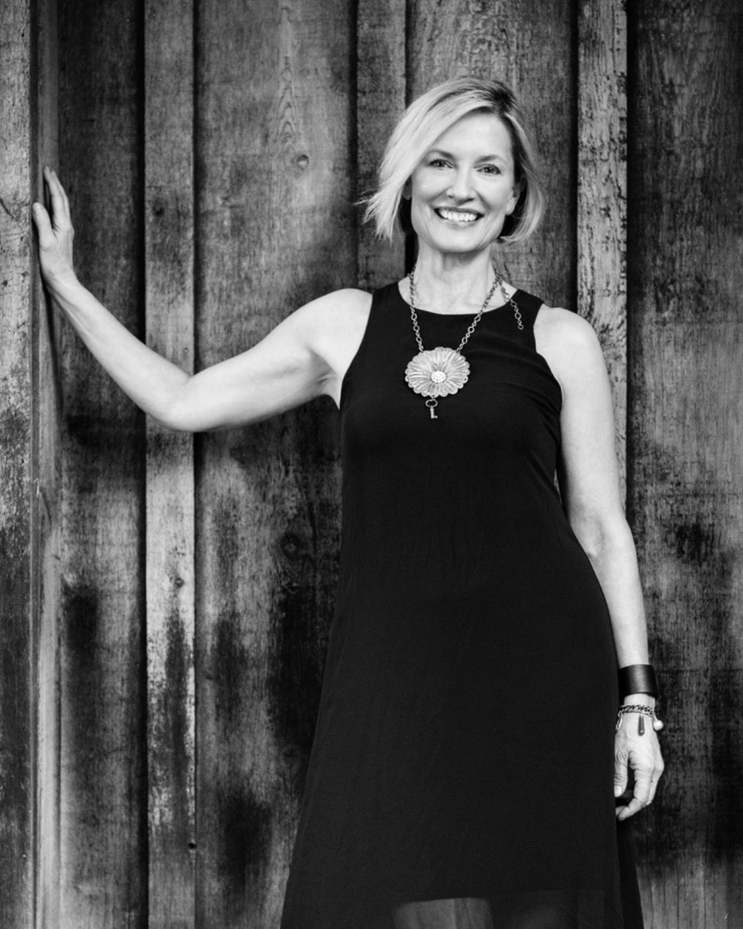 Black and white photo of a woman with short blonde hair smiling, wearing a sleeveless black dress with a large floral necklace, standing against a wooden wall, her left hand touching the wall.