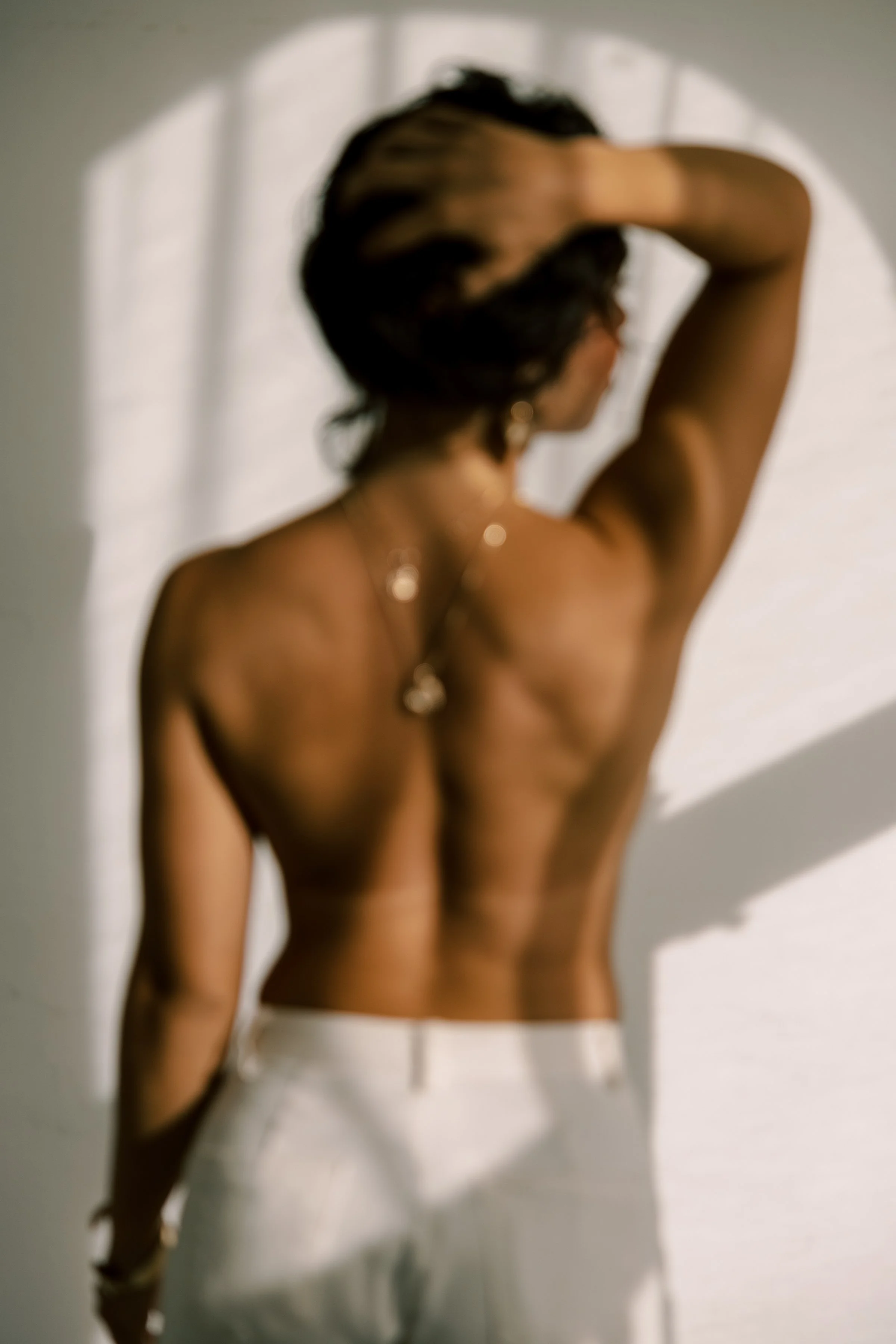A woman with tan skin and dark hair, touching her hair and facing away, wearing earrings and white pants, standing in front of a white wall with shadows.