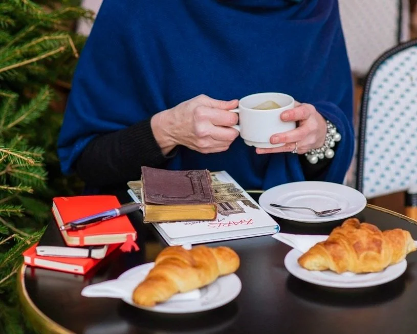 Person holding a cup of coffee or tea, sitting at a table with croissants, a small plate, and a pencil, with Christmas decorations in the background.