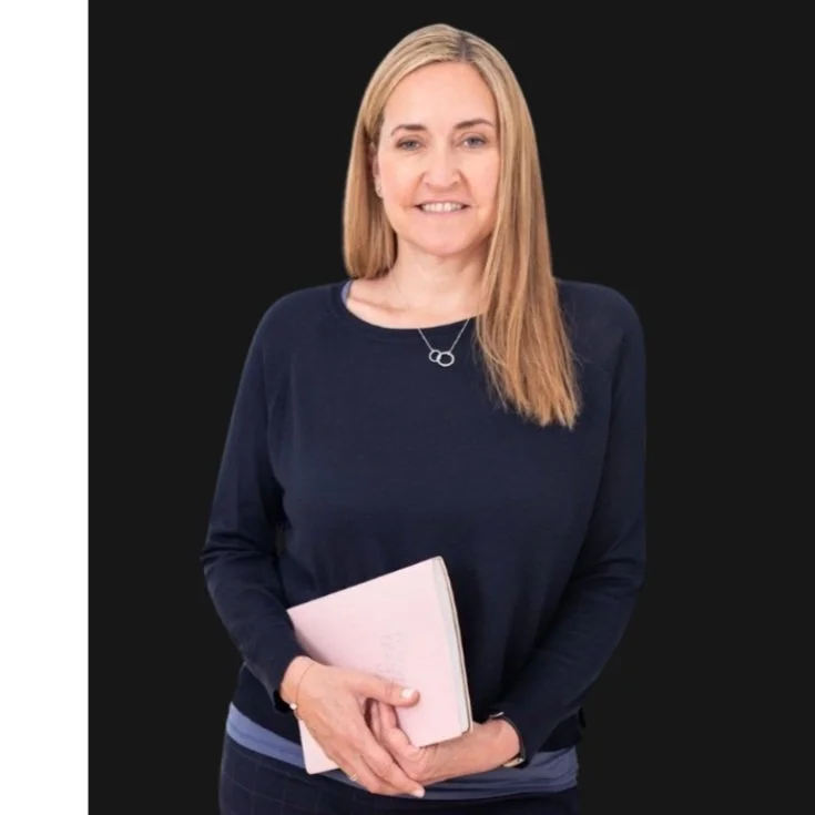 A woman with long blonde hair, wearing a dark long-sleeved top, holding a pink notebook, standing against a black background.