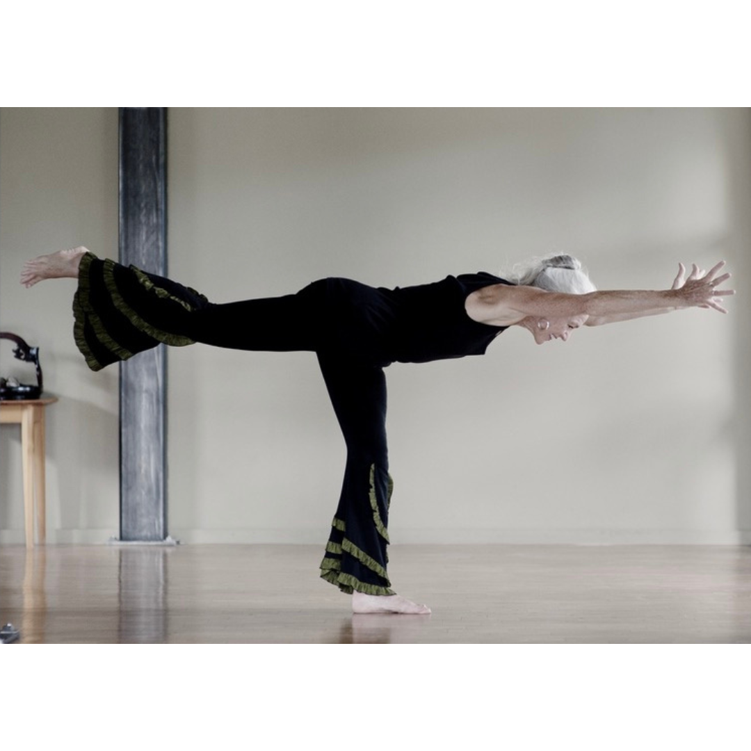 An elderly woman practicing yoga in a studio, standing on one leg with her other leg extended backward and her arms stretched forward.