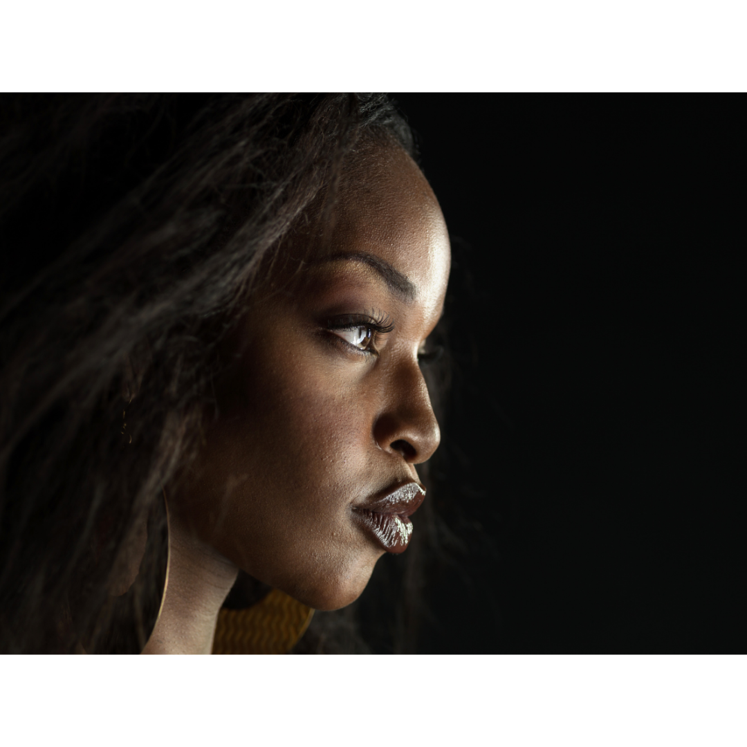 Close-up side profile of a woman with dark curly hair, golden skin, and dark makeup against a black background.