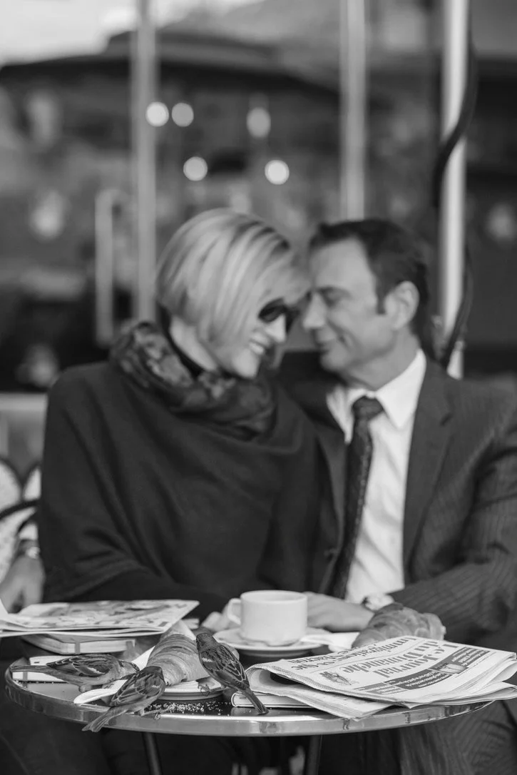 A black-and-white photo of a man and woman smiling and leaning close together at a table in a café or restaurant. There are newspapers, a coffee cup, and some bread on the table. The background shows blurred lights and structures, suggesting an outdoor or urban setting.