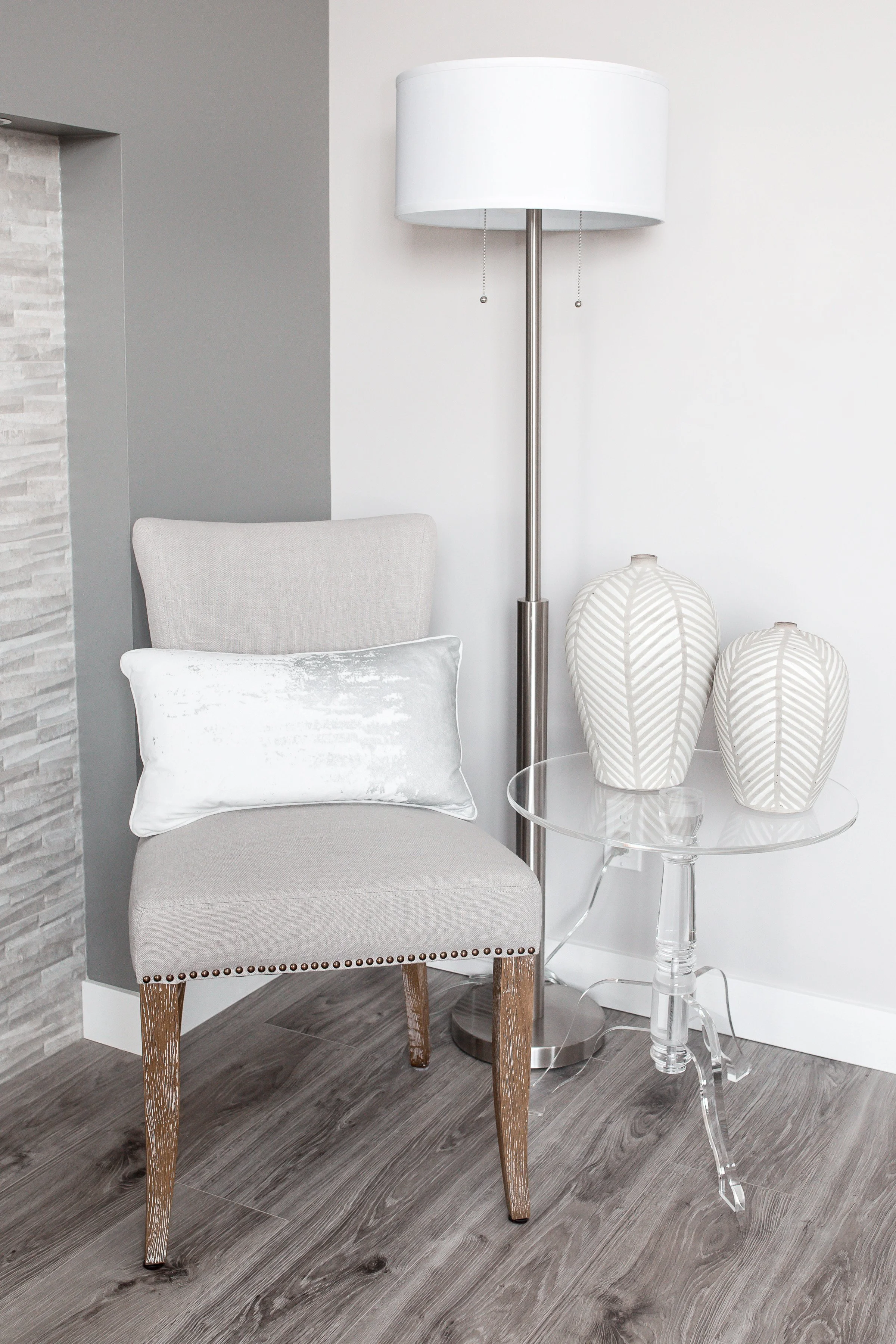 A beige upholstered chair with wooden legs, a white throw pillow, a tall white floor lamp, a clear acrylic side table, and two white decorative vases with leaf patterns on a wooden floor against a white and gray wall.