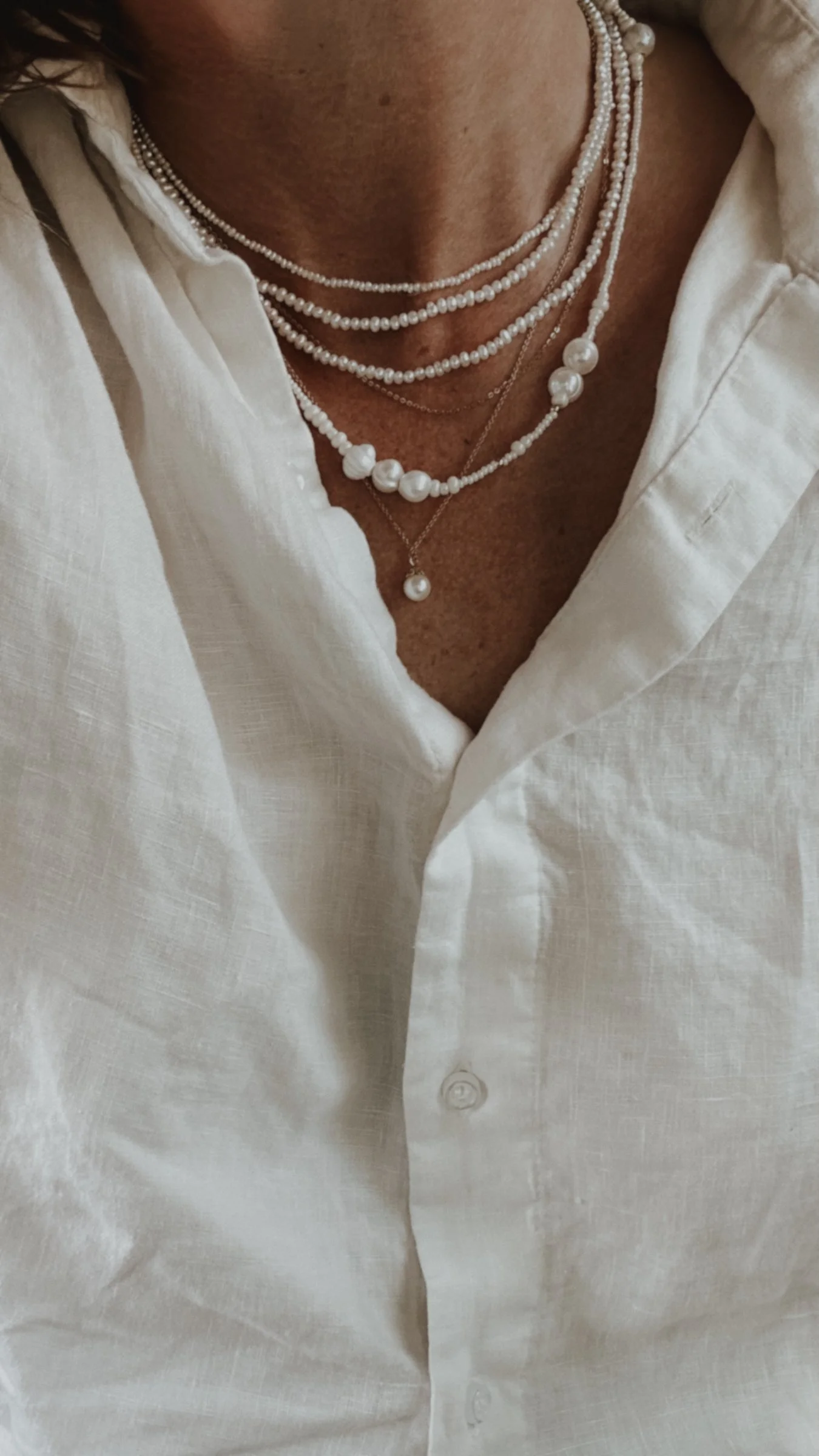 Close-up of a woman's chest wearing layered pearl necklaces and a white button-up shirt.