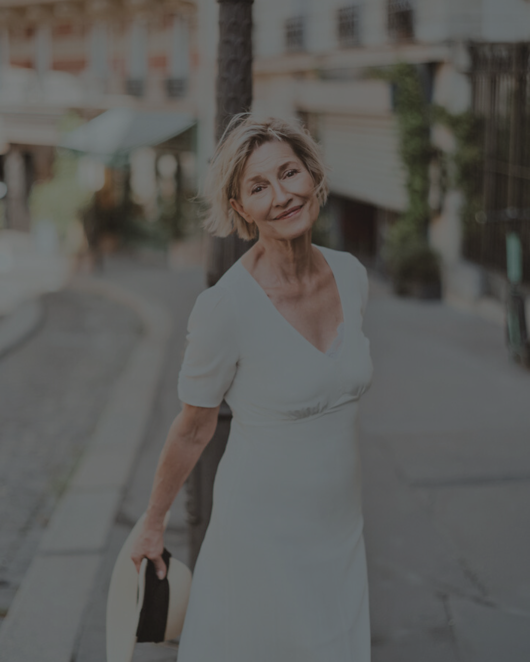 An elderly woman with short, blonde hair wearing a white dress, standing on a sidewalk in an urban setting with residential buildings in the background.