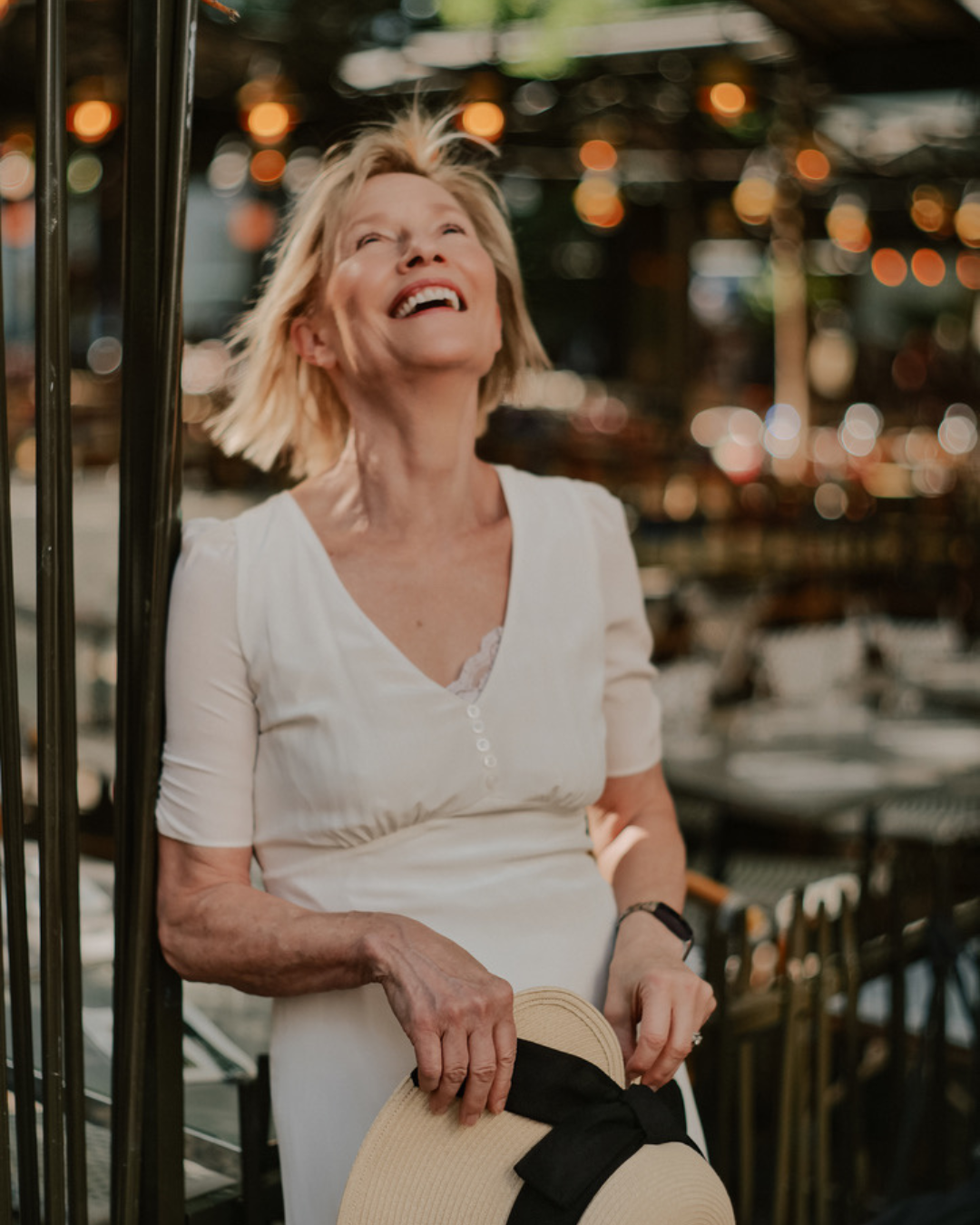 A joyful woman with short blonde hair, wearing a white dress, holding a wide-brimmed hat with a black ribbon, standing in a lively outdoor setting with warm bokeh lights in the background.
