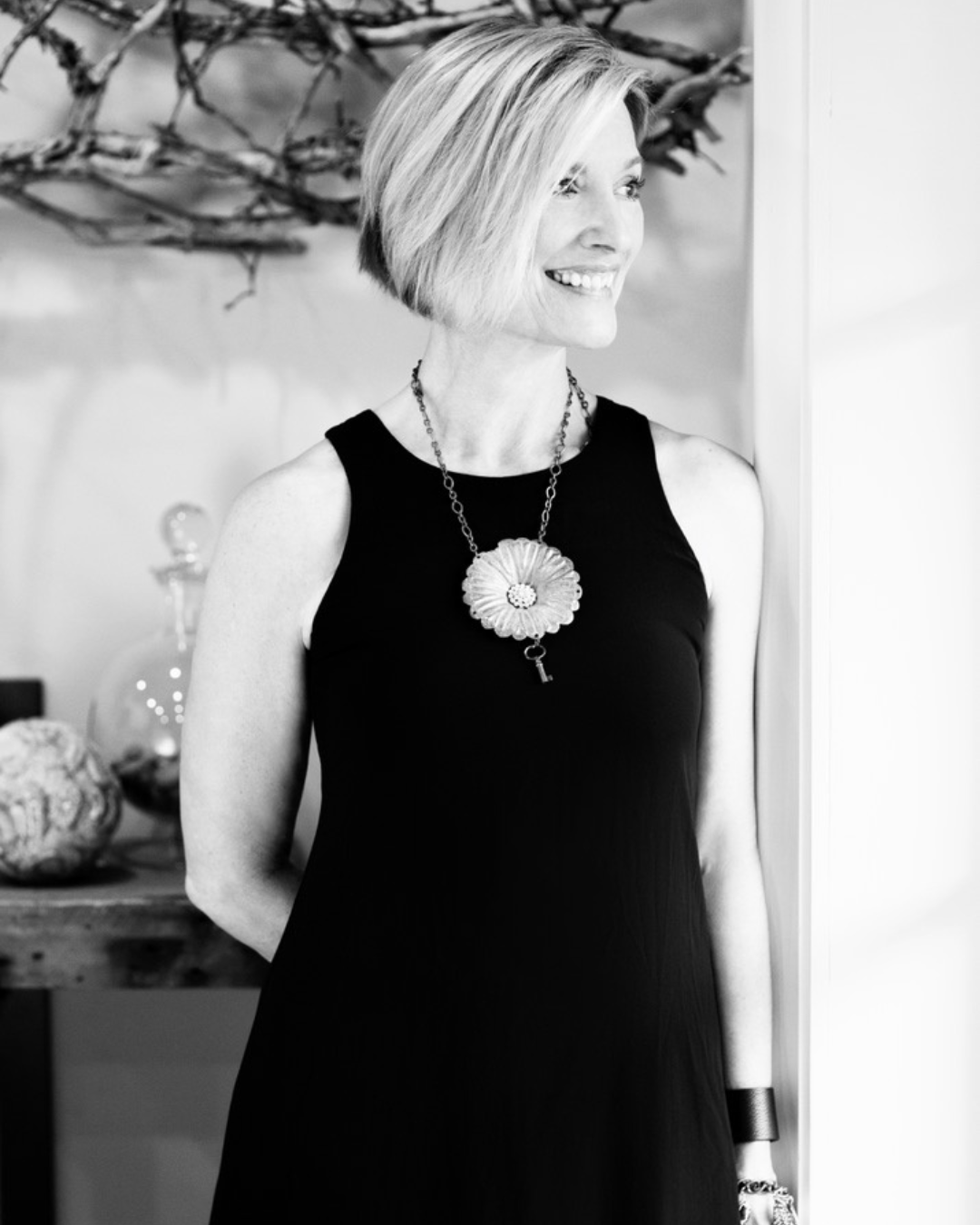 A smiling woman with short blonde hair in a black sleeveless dress, wearing a large flower-shaped necklace, standing indoors with a decorative branch arrangement in the background.