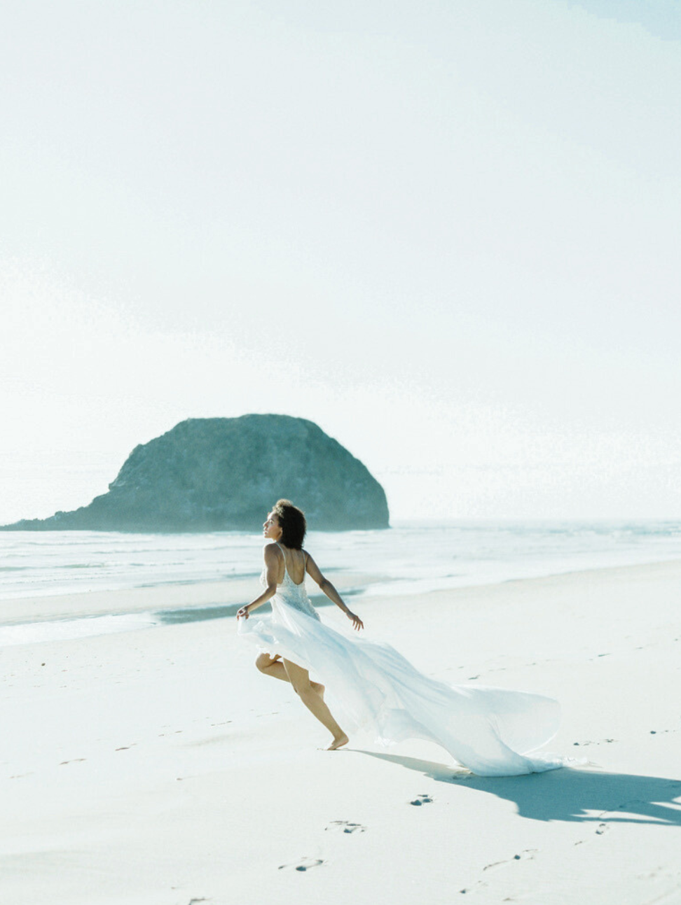 A woman in a white dress running on a sandy beach with a large island or rock formation in the background.